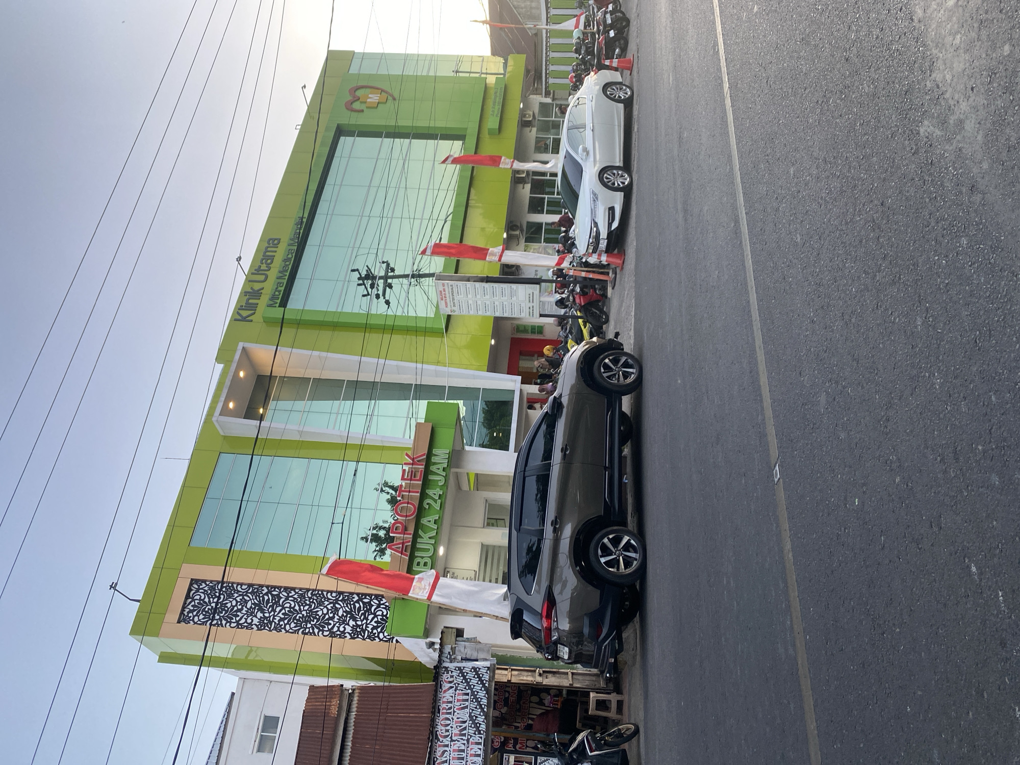 The image captures the exterior of a modern, multi-story building identified as "Klinik Utama Mitra Medica Mandiri" and an adjacent "Apotek Buka 24 Jam" (Pharmacy Open 24 Hours) in Bantaeng, Indonesia. The building features a bright green facade with large, reflective glass windows. Decorative elements include a patterned panel on the upper left and a logo incorporating a red heart with an "M" inside on the upper right. Several Indonesian flags, red and white, are prominently displayed.
In the foreground, a dark grey SUV is parked on the right side of the street, facing left. A white sedan is parked on the left side of the street, also facing left. A busy street with a yellow line marking the lane is visible in front of the vehicles. Various motorcycles are parked in front of the pharmacy and clinic. Several individuals, some wearing helmets, are standing or walking near the entrance of the pharmacy and clinic. To the far left, a food stall with an awning displays signs that appear to be related to food sales. The sky is clear and bright, indicating daytime. The overall scene depicts a commercial area in the city of Bantaeng.