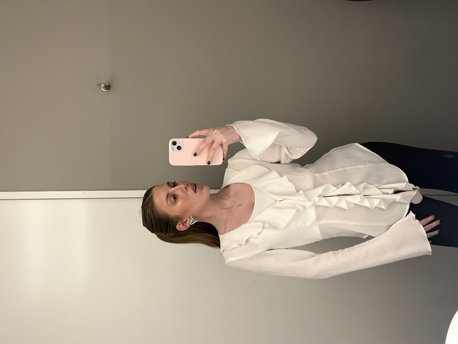 A woman in black leggings and a white, frilly shirt takes a selfie in what appears to be a dressing room. She holds a pink phone case, with black nail polish visible on her fingers. Her hair is pulled back, and she wears triangular earrings. The room features a gray upper wall and a white lower section. A doorknob is also visible on the upper wall.