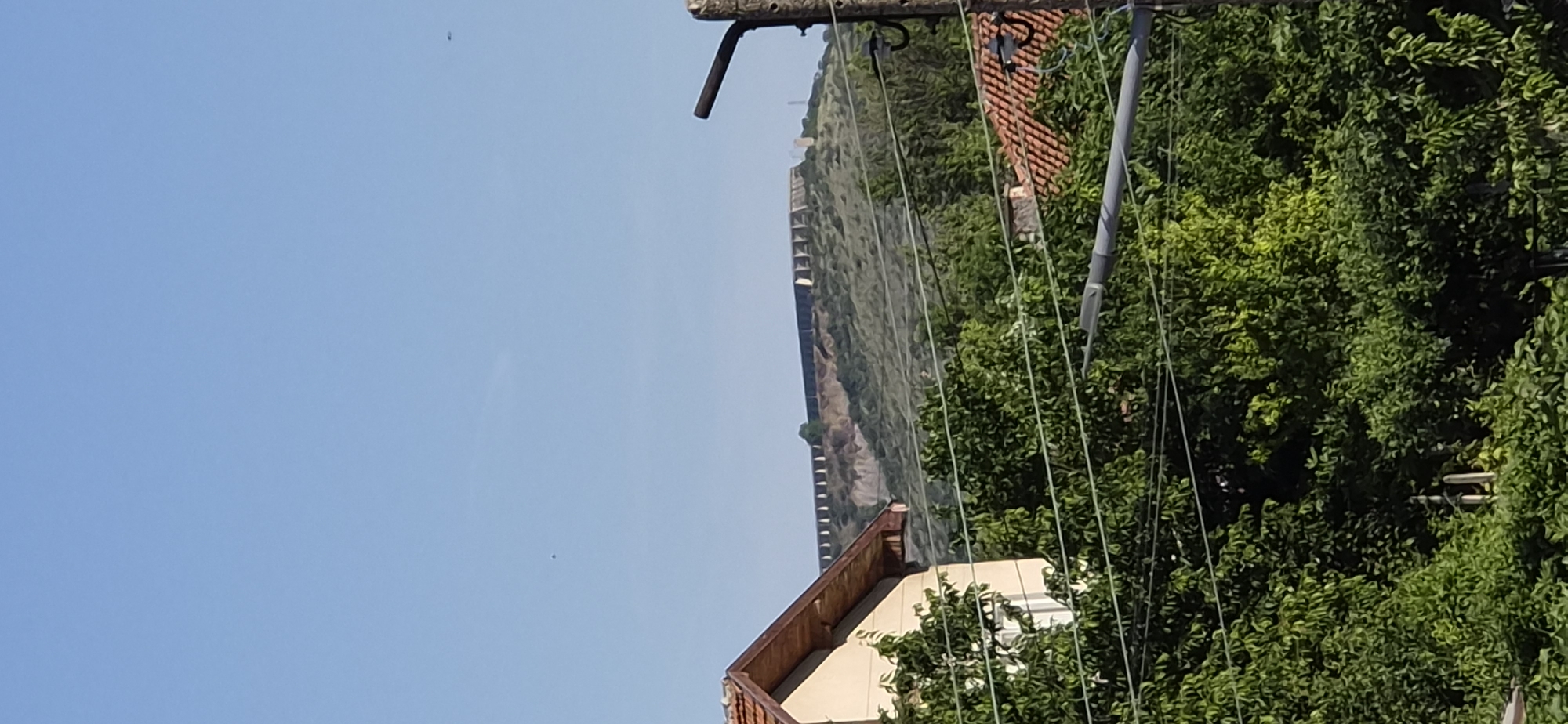 A bright, clear, sunny day is depicted in this vertical outdoor photograph. In the foreground, lush green trees fill the bottom half of the image, with the peak of a residential house with a reddish-brown tiled roof visible on the left side. Power lines are strung across the frame, supported by a rough, wooden pole on the far right. In the background, a long, imposing stone wall stretches along the crest of a grassy, undulating hill. The wall appears ancient and weathered, with a series of buttresses or supports visible along its length. The sky is a clear, uninterrupted blue, with a couple of distant, dark specks that could be birds or insects. The overall scene suggests a rural or semi-rural setting, with a prominent historical landmark situated in the distance. This image likely depicts a view of the Radilovo Fortress, a historical site located in Radilovo, Bulgaria.
