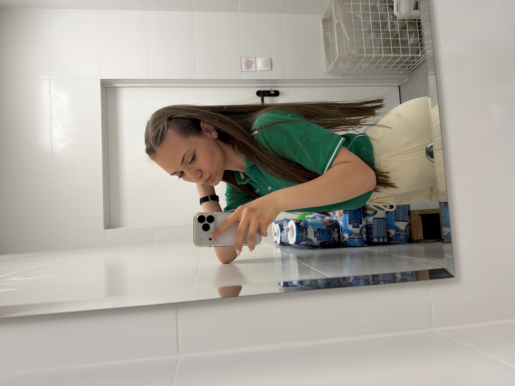 A young woman is taking a selfie in a bathroom mirror. She is wearing a green polo shirt with white trim and light-colored pants. Her hair is long and brown. In the mirror's reflection, behind her, are several rolls of toilet paper. The bathroom has white tile walls and a white wire basket on the wall. The location is potentially in Sofia, Bulgaria.