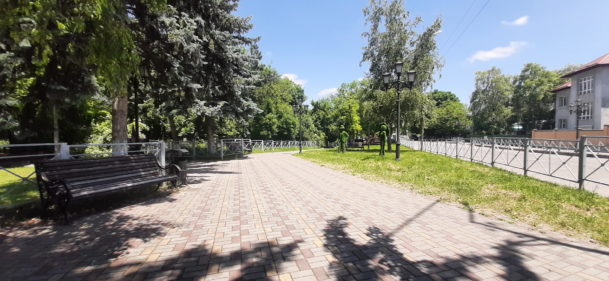 The image depicts a sunlit park pathway in Yessentukskaya, Russia. The pathway is paved with light-colored rectangular bricks laid in a herringbone pattern. To the left of the pathway, a dark wooden park bench with metal legs is situated on a grassy area bordered by a decorative metal fence. Beyond the bench and fence, large evergreen trees with dense foliage are visible. Further down the path, more trees, streetlights, and two topiary figures, resembling people, are present on a grassy area also enclosed by a metal fence. To the right of the pathway, the metal fence continues, separating the park from a street or road. A building with light-colored walls and multiple windows is partially visible in the background on the right. The sky is blue with scattered white clouds. The scene is devoid of people actively engaged in any actions.