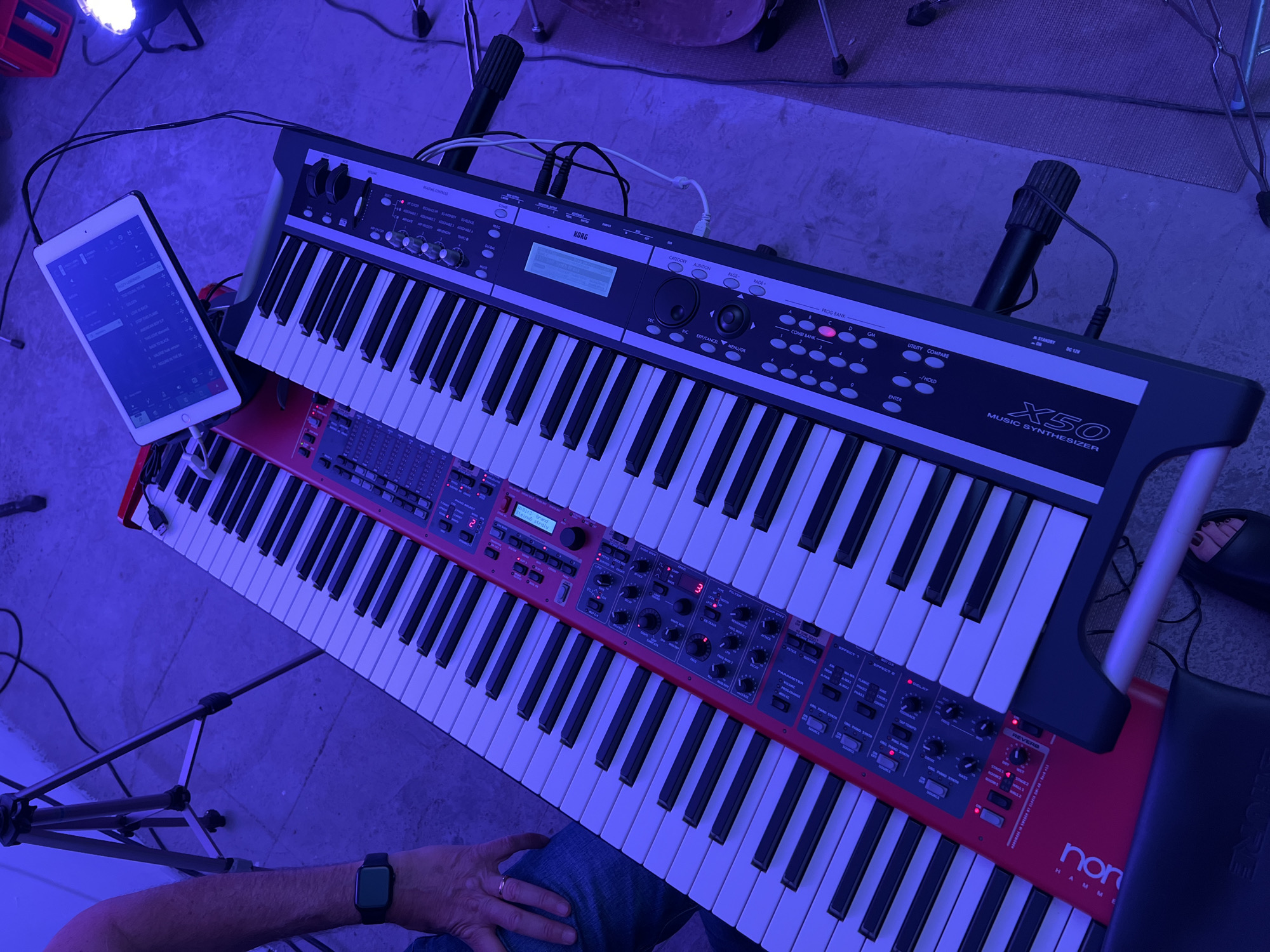 The image depicts a musical setup within an indoor environment in Santeramo in Colle, Italy, illuminated primarily by a strong blue light, giving the scene a monochromatic appearance. Two electronic keyboards are central to the composition. The lower keyboard is a red Nord Stage 3 (identifiable by the "Nord" logo and "HANDMADE IN SWEDEN BY CLAVIA DMI AB" text), featuring 73 white and black keys. Positioned above and to the right of the Nord keyboard is a black and grey Korg X50 Music Synthesizer, discernible by its "KORG" and "X50 MUSIC SYNTHESIZER" branding, with 61 white and black keys.
An iPad with a white bezel is elevated on a stand or directly on the Nord keyboard, displaying a digital interface. Multiple black and white cables connect the keyboards and iPad to unseen peripherals. To the upper left, a metallic tripod stand is visible.
A person's left arm and hand are visible in the lower-left quadrant. The individual wears a dark smartwatch with a green display on their wrist and a gold ring on the fourth finger of their left hand. The person is wearing blue denim jeans. A foot, clad in a dark sandal or flip-flop, is partially visible in the lower right corner, adjacent to a metallic stand component.
The floor is a light-colored, possibly concrete surface. A partially visible black object with the word "SHURE" is present in the bottom left. A black microphone stand extends from the right, beneath the Korg keyboard. Another dark, indeterminate object, possibly part of a drum or instrument, is visible on the far right. The overall setting suggests a musical performance or practice space.