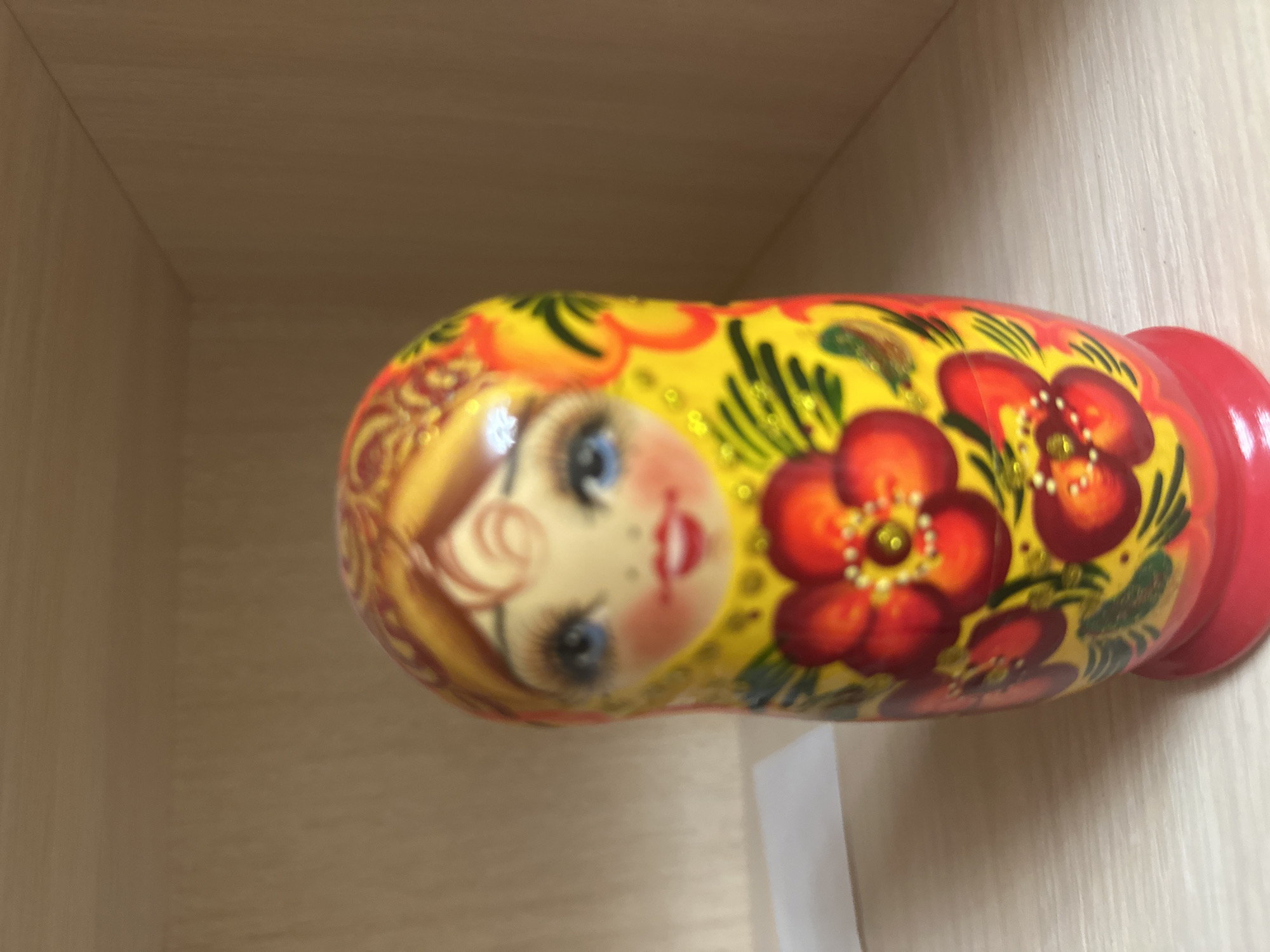 A matryoshka doll, also known as a Russian nesting doll, sits on a wooden shelf. The doll is predominantly yellow and red with a painted face featuring blue eyes, rosy cheeks, and red lips. It is decorated with floral motifs in shades of red and green, with hints of yellow and gold. The doll is positioned in the center of the image, with its base resting on the shelf. The background consists of a light brown wooden shelf. The image does not provide any information about its location in Kazan, Russia.