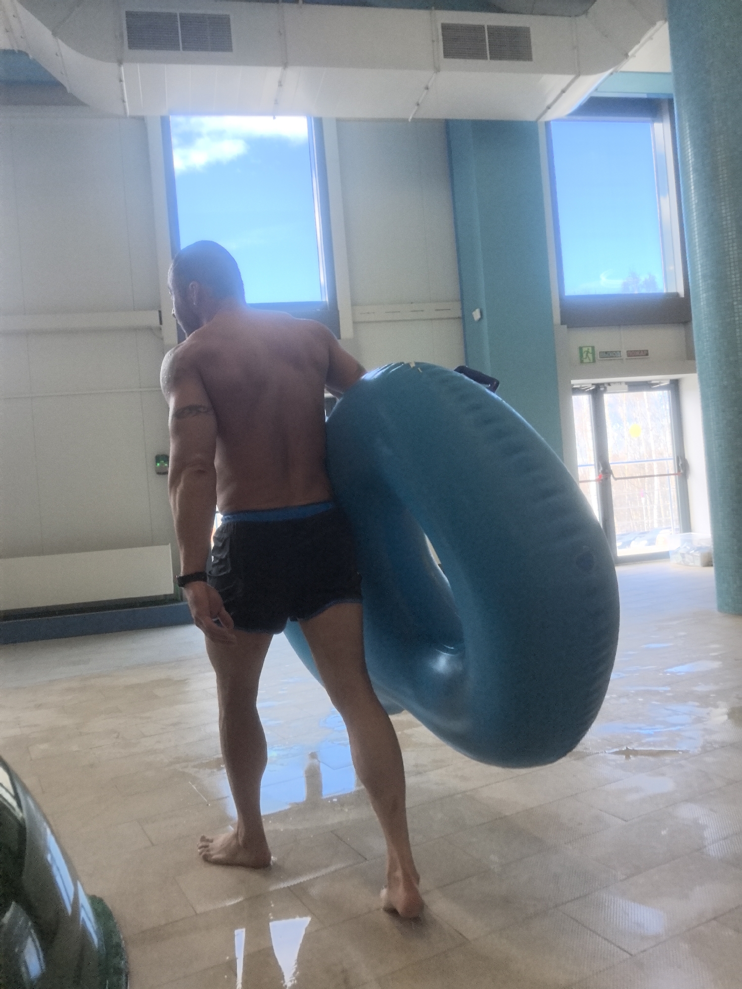 A shirtless man in black shorts holds a large blue inflatable tube in a water park-like setting. The man is seen from behind, and he is barefoot. The floor is tiled and wet. There are large windows showing a blue sky outside. In the background, there's a doorway with an exit sign in Russian. The water park's design includes light blue and white walls. Given the Russian signage and architectural style, the location is likely in Nizhny Novgorod, Russia.
