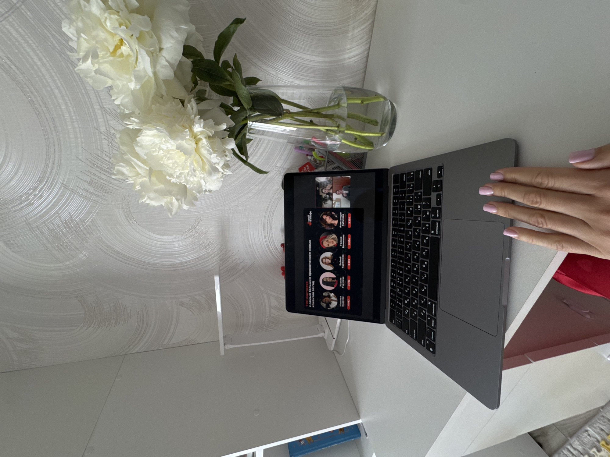 Here is a detailed description of the image:
The image appears to be taken from an overhead perspective of a workspace. On a white desk, there's a laptop displaying a screen with the text "ТОП партнеров" and names in Russian, indicating a list or ranking of partners with client attraction metrics. A vase with white flowers is placed beside the laptop. A small desk lamp is positioned to the left of the laptop. A hand with manicured nails is partially visible at the right edge of the desk. The background includes a textured, possibly wallpapered, surface and the edges of a white shelf with a glimpse of a blue object on it. The environment suggests a personal office or home setting.
It is not possible to determine the location in Sochi, Russia, from the given image.