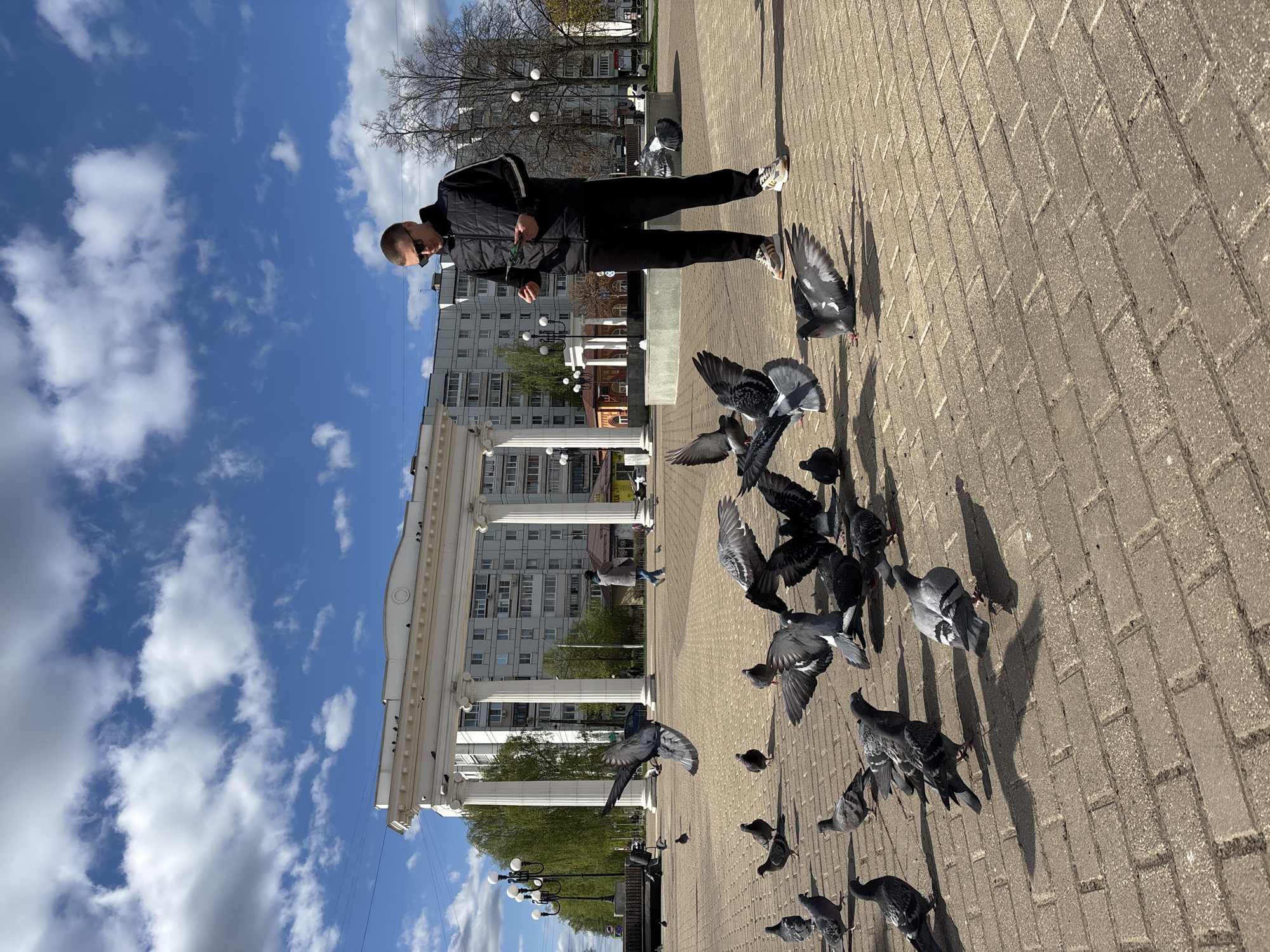 A man in black clothing feeds pigeons on a brick-paved square. The location is Chaikovsky, Russia, evidenced by the architecture. The man stands amid a flock of pigeons, some in flight, others on the ground. In the background is a white colonnade in front of a building, along with street lights and trees. The sky is blue with scattered clouds.