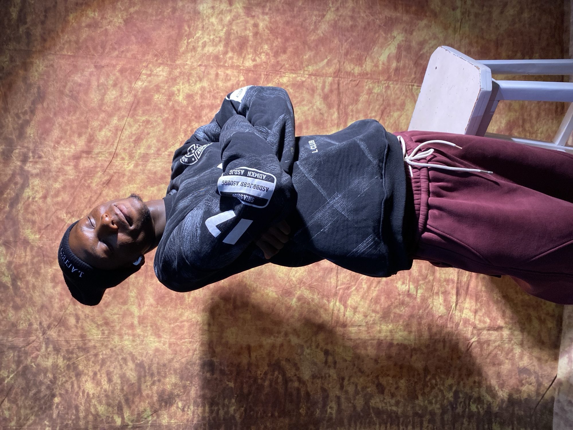 A young Black male stands with his arms crossed, facing slightly to the right, his eyes closed and a relaxed smile on his face. He is wearing a black beanie with white lettering that reads "JAMES", a black sweatshirt with white graphic elements on the sleeves and across the chest, and maroon sweatpants. He has white earbuds in his ears. He is standing in front of a mottled brown and orange backdrop. To his right, partially visible, is a small, white stool with a worn wooden top. The lighting casts a shadow across the backdrop and highlights the subject. The overall scene appears to be a casual studio portrait. There is no visible indication of the location within the city of Barakin Akawo, Nigeria.
