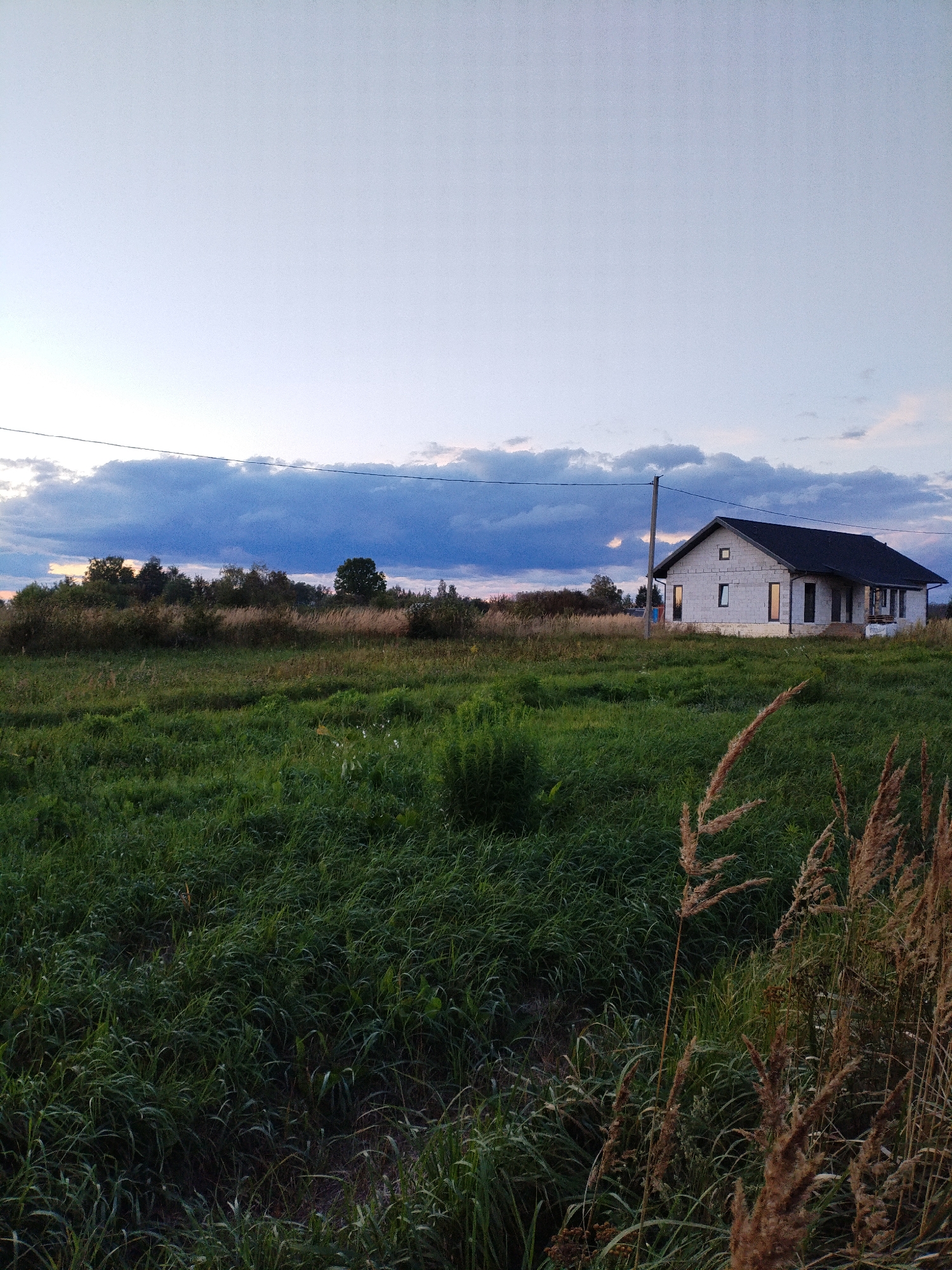The image depicts a partially constructed house in a rural setting near Bialynichy, Belarus. The house, made of white cinder blocks with a dark roof, has several windows and doors. It is situated in a grassy field with some dry grass in the foreground. In the background, there are trees and a cloudy sky. A utility pole and wire are visible in the mid-ground, running horizontally across the image. The scene appears to be captured at dusk, with the sky showing a gradient of light blue and grey.