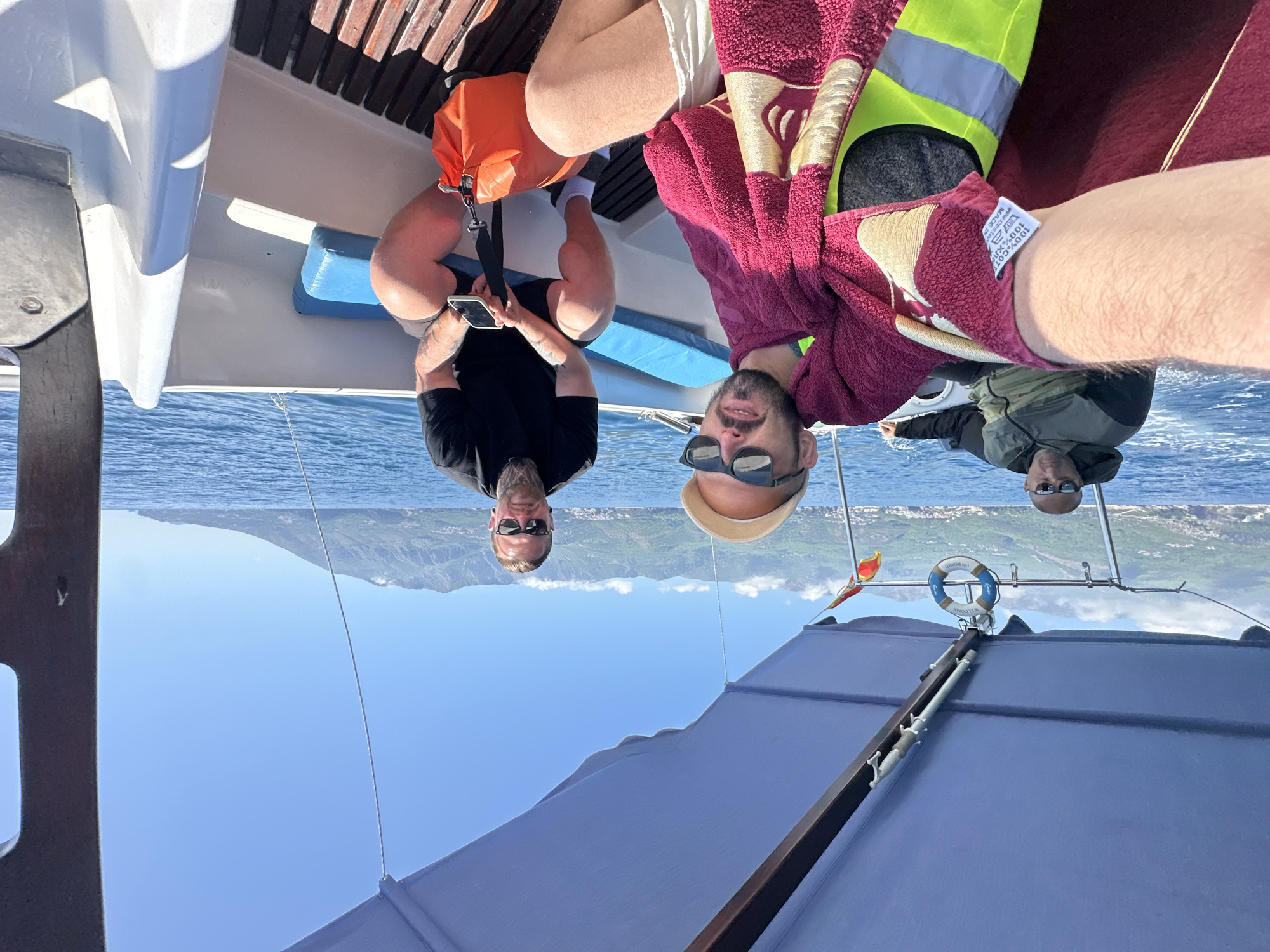 The scene is set on a boat at sea, possibly near Budva, Montenegro given the mountainous coastline in the distance. Three men are visible: one is sitting wearing sunglasses and a black t-shirt, looking at a phone, with an orange bag next to him. Another man is wearing sunglasses and is wrapped in a maroon towel and a yellow safety vest. A third man with sunglasses is wearing a green jacket. A life preserver ring is visible on the boat. The sky is clear and blue.