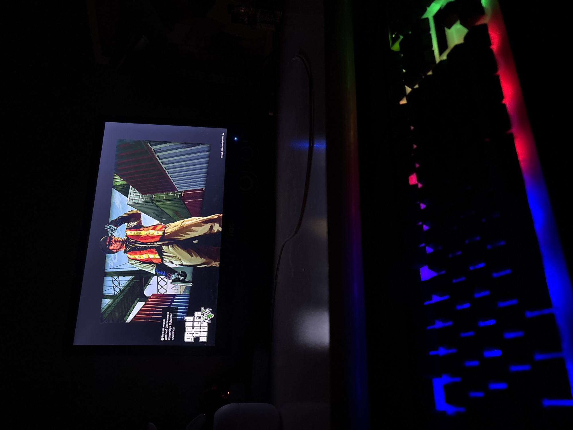 The image shows a dark room with a monitor displaying the loading screen of Grand Theft Auto V. The loading screen depicts a construction worker standing in front of stacked shipping containers. To the right of the monitor, a gaming keyboard with RGB backlighting is visible, with the colors shifting from green at the top to blue at the bottom. The background is dark, suggesting a poorly lit room. The presence of gaming peripherals implies that this is likely a home gaming setup. There is no information in the image to determine a specific location in Yerevan, Armenia.