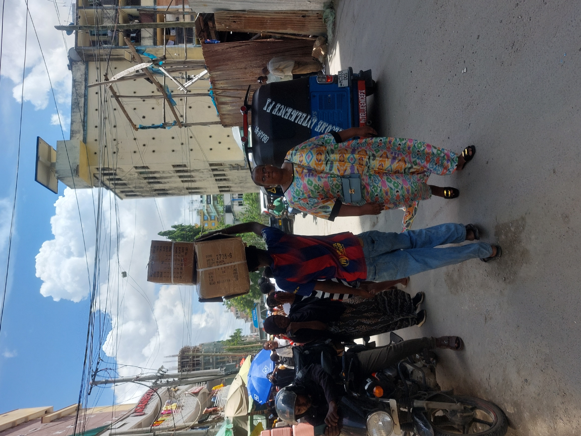 This vibrant daytime street scene in Dar es-Salaam, Tanzania, captures various aspects of urban life. The setting features a paved street flanked by multi-story buildings, some appearing aged or under construction, against a bright blue sky dotted with fluffy white clouds.
In the foreground, several individuals are engaged in daily activities. A woman in a colorful, patterned dress and golden sandals, carrying a light blue shoulder bag, looks directly towards the camera with a neutral expression. Nearby, a man expertly balances two stacked cardboard boxes on his head, a common method of transporting goods. He wears a red and blue striped shirt, possibly a football jersey, with a visible "UNHCR" logo, and blue jeans. Other people are seen walking or standing, including one person on a black motorcycle wearing a helmet, and another person in a black and white patterned skirt.
Vehicles include a blue and black Bajaj Re rickshaw (tuktuk) parked by the side of the road. Visible text on the back of the rickshaw includes "WELCOME INTELLIGENCE FX", "OBAH-89", and license plate details "MC333 DQS". The cardboard boxes carried by the man have labels such as "ART NO: 2735-6", "COLOR: BLACK", "QTY: 14 PRS", "MEAS: 62x25x34 CM", and "0085". The scene conveys a sense of bustling activity typical of a busy city street, with everyday commuting and commerce taking place under clear, sunny weather.