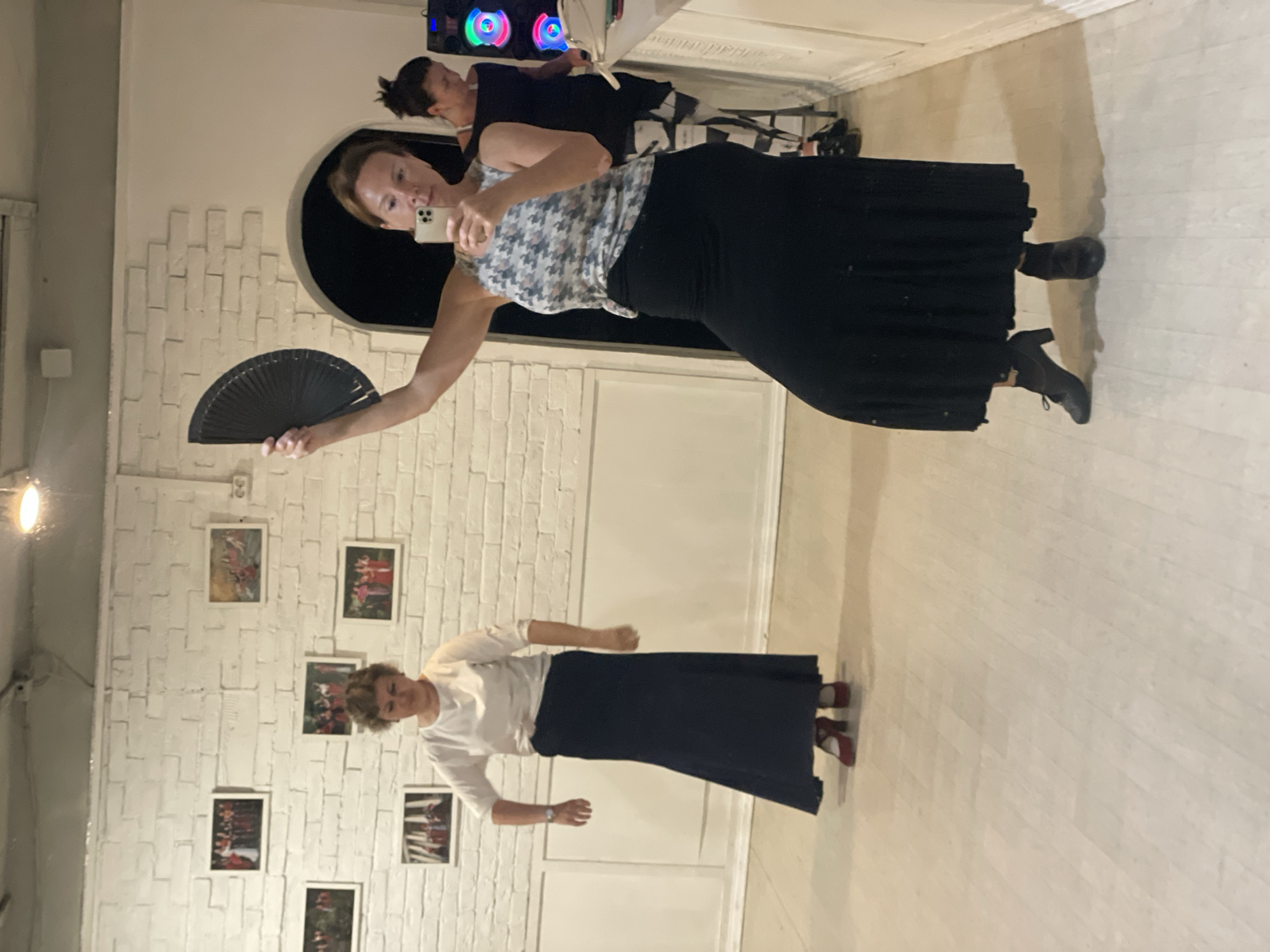 In a dance studio located in Sochi, Russia, two women are captured in a mirror selfie. The woman in the foreground, in a black and white patterned sleeveless top and a long black skirt, holds a black fan in her right hand and a smartphone in her left, taking the photo. She is wearing dark heeled boots. Behind her, a second woman in a white long-sleeved shirt and a long dark blue skirt stands with her hands down, wearing red heeled shoes. The studio has white brick walls adorned with framed pictures of what appear to be dancers. The floor is a light-colored wood. To the right, a colorful speaker system is visible.