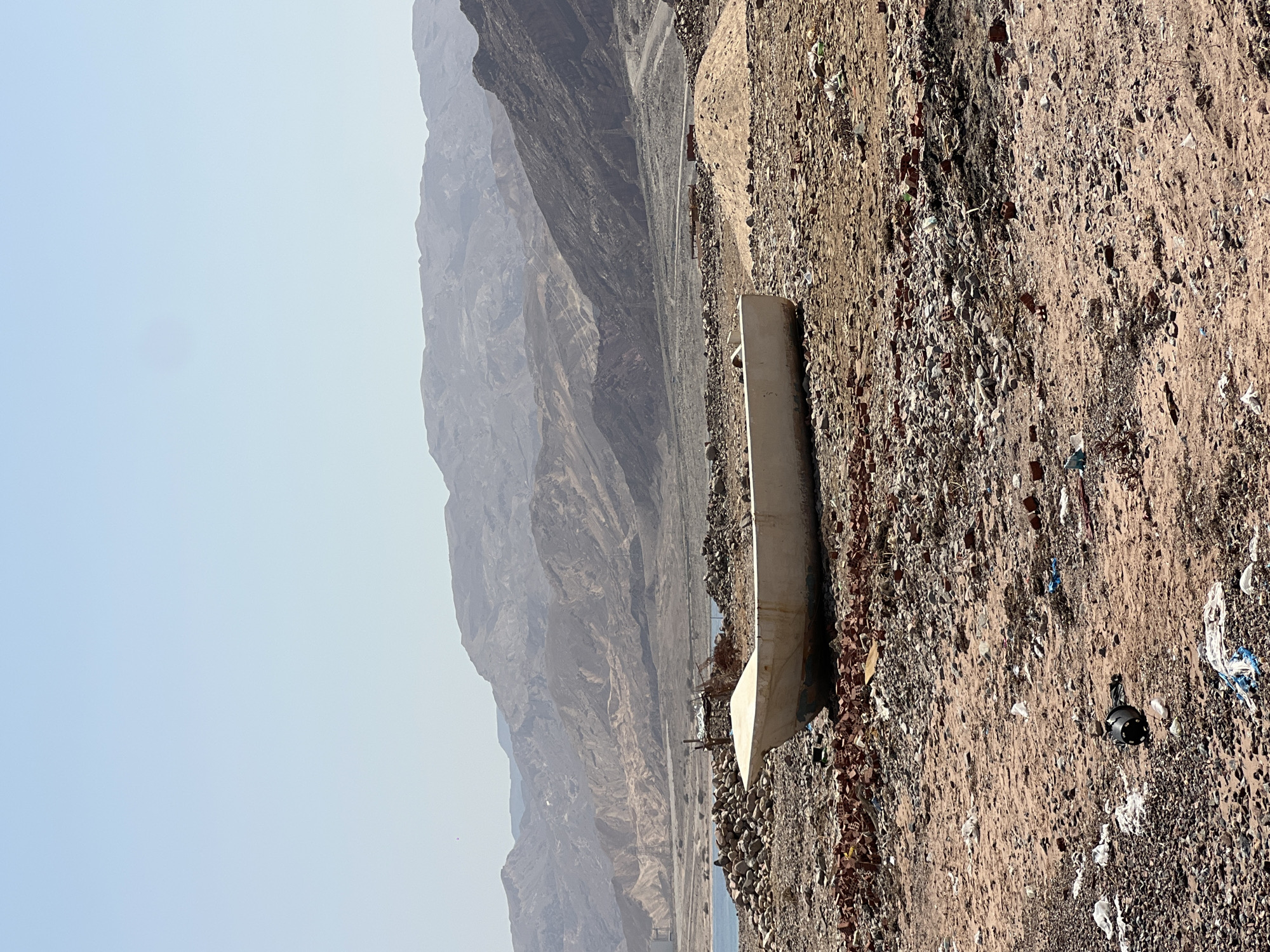 An abandoned white boat lies beached on a debris-strewn, sandy shore with a rocky coastline. In the background, rugged, layered mountains rise under a clear, pale blue sky. The ground is covered with various sizes of rocks, broken red bricks, and scattered white plastic debris. The water is visible in the distance to the left. The scene appears to be a desolate and neglected coastal area in Egypt.