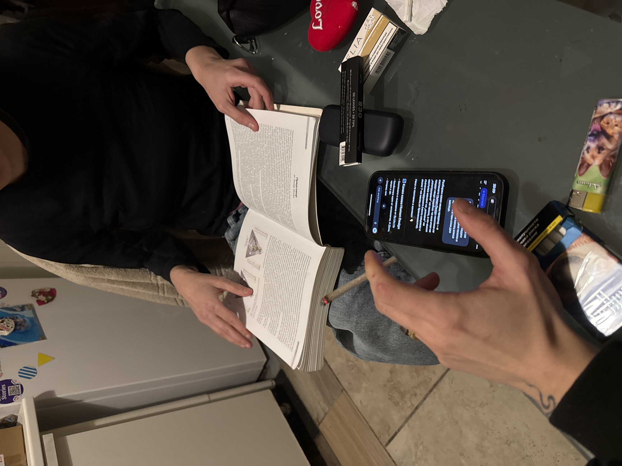 Here's a detailed description of the image:
**Scene Overview:**
The image presents an indoor setting, possibly a kitchen or living area. The viewpoint is an overhead shot. The scene depicts a person seated at a table, engrossed in reading a book. Another person is visible, seemingly capturing an image of the scene with their phone.
**People:**
* **Person 1 (Reader):** Dressed in a black long-sleeved shirt, their hands are actively engaged in holding open a thick book.
* **Person 2 (Photographer):** Only their hand and part of the arm are visible. Their hand is holding a smartphone, suggesting they are taking a photo or recording a video of the scene. A tattoo is visible on their wrist.
**Objects:**
* **Book:** A large book filled with text is open.
* **Smartphone:** An iPhone screen displays a messaging app with texts written in Bulgarian.
* **Rolling tobacco items:** A pack of cigarette rolling papers are placed on the table, along with a pack of IQOS heatsticks, an empty pack of OCB tips, and a lighter. There is a lit cigarette.
* **Other items on the table:** A small red heart-shaped decoration and a black pouch.
* **Refrigerator:** A white refrigerator is visible in the background, adorned with magnets and stickers.
**Actions and Interactions:**
* Person 1 is reading a book.
* Person 2 is using their phone, possibly to take a picture or record a video of Person 1 reading. The cigarette is held in their left hand.
**Environment:**
* The scene takes place indoors.
* There is a table.
* There is a white refrigerator in the background.
**Location:**
The messages on the phone screen are written in Bulgarian. The book is also written in Bulgarian. This would place the setting in Bulgaria. More precisely, Sofia is written on the lighter that is visible, so that would place the location of the image in the city of Sofia in Bulgaria.