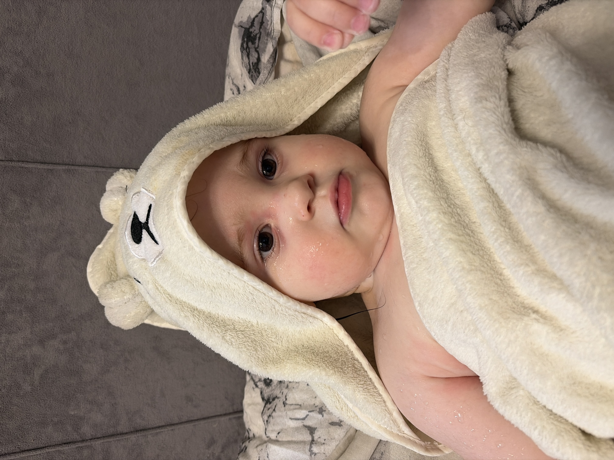 A baby with damp hair and a few droplets of water on their face is wrapped in a hooded towel with bear ear appliques. The background consists of a gray, seemingly textured surface. The scene is likely indoors. The precise location within the city of Chaikovsky, Russia, cannot be determined from this image.