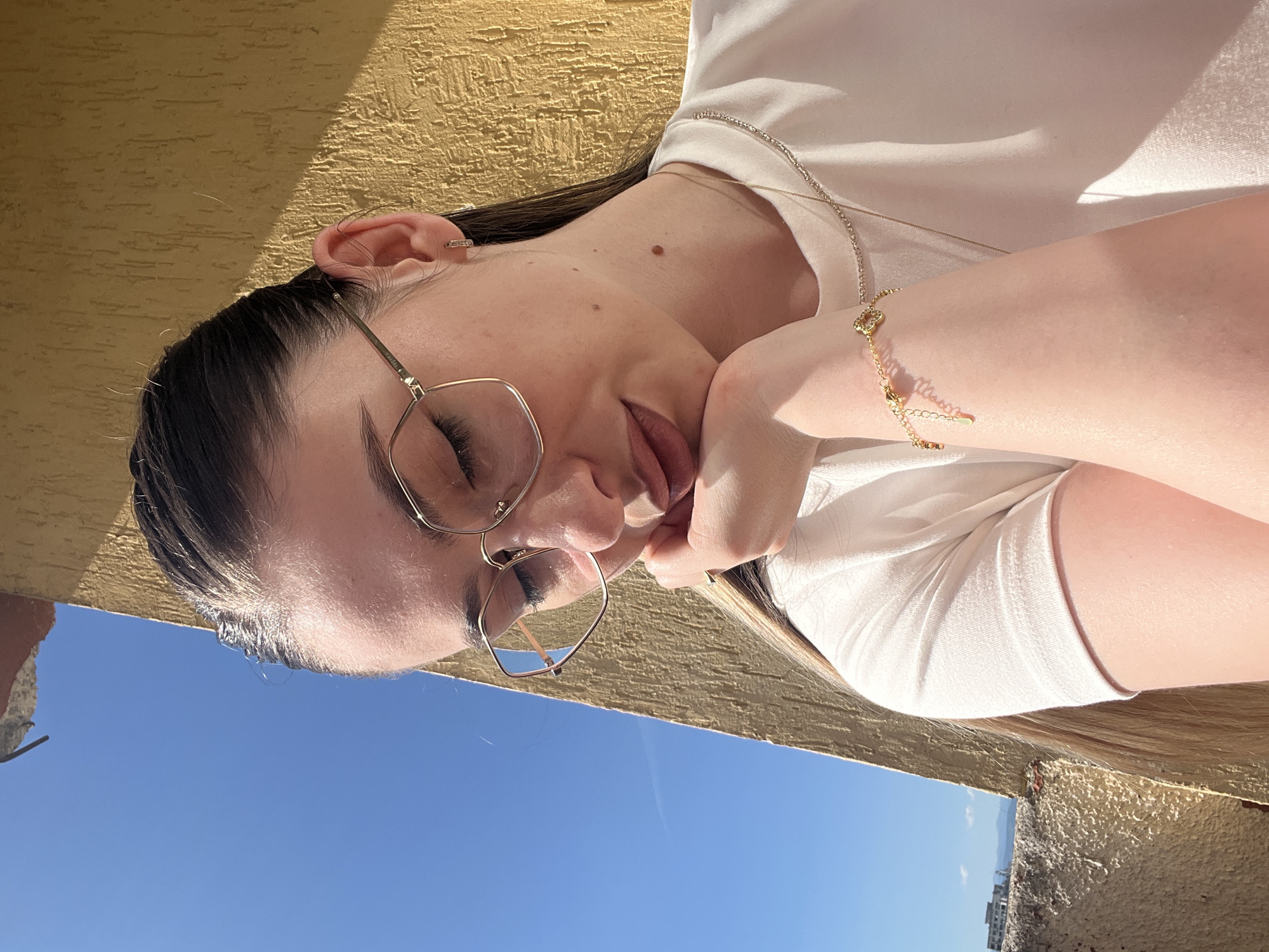A young woman with her eyes closed and her head resting on her hand is captured in a close-up shot against a bright blue sky and a textured yellow wall. She is wearing large, gold-rimmed glasses and a white t-shirt. A delicate gold necklace and a bracelet with a heart charm adorn her neck and wrist. Her dark hair is pulled back, and sunlight illuminates her face and right arm. The background suggests an outdoor urban setting, possibly a rooftop or balcony, with glimpses of distant buildings and mountains.