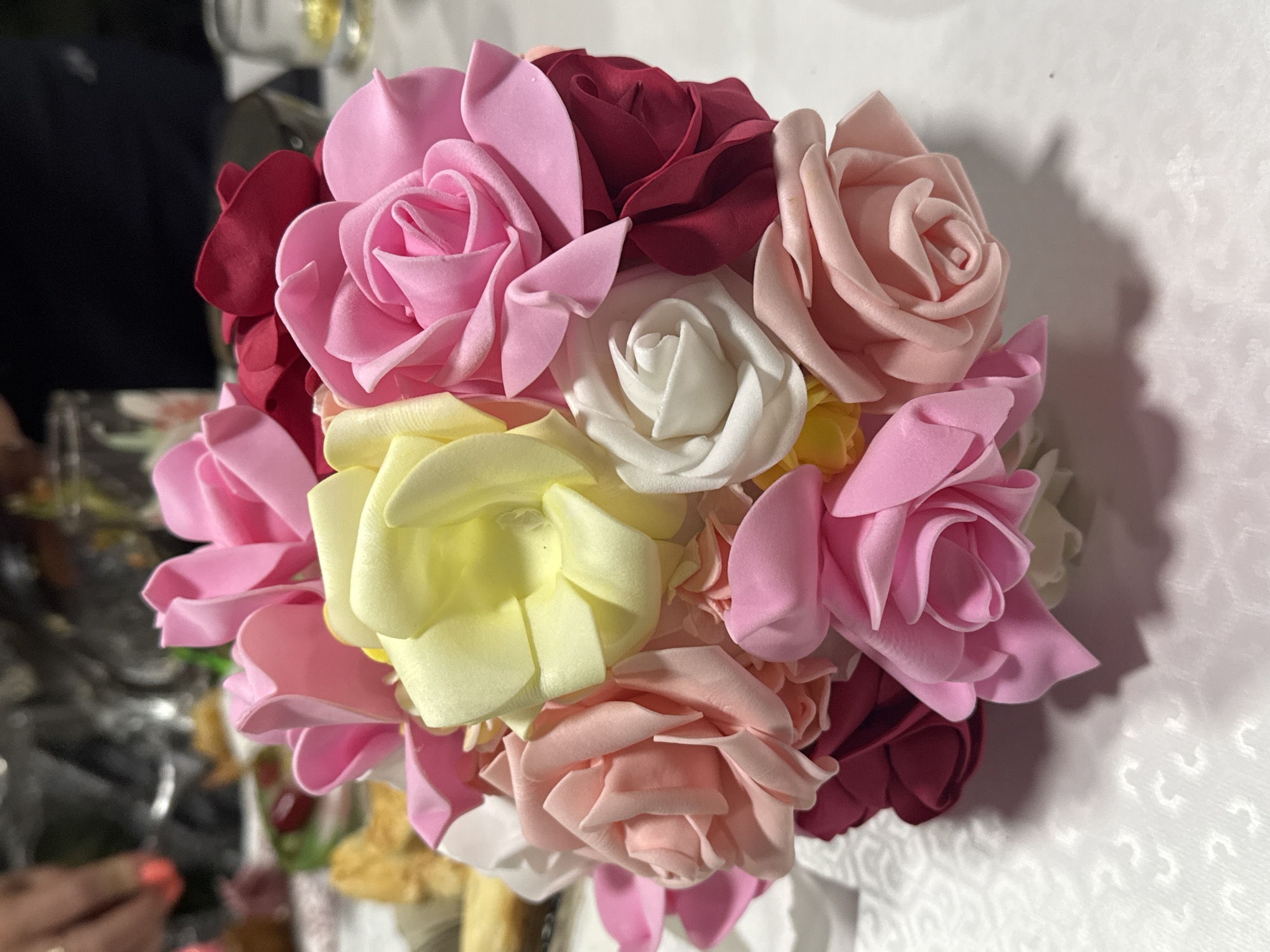A bouquet of artificial roses in various shades of pink, white, yellow, and deep red is arranged on a white, textured tablecloth. The bouquet is the central focus of the image. In the blurred background, hints of a celebratory setting are visible, including what appears to be a person's hand with manicured nails in the upper left corner, and scattered food items on plates. The lighting suggests an indoor environment, possibly a dining area. The image does not contain information specific to Tash-Moinok, Kyrgyzstan.