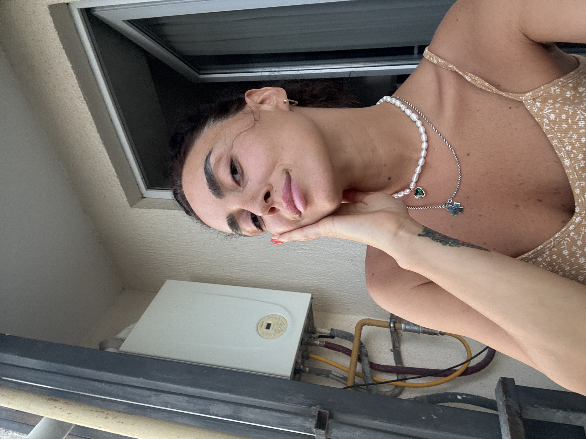 A woman is captured in a selfie taken outdoors, likely on a balcony, given the presence of a water heater and pipes on the wall behind her. She is leaning her head on her hand, with her right cheek resting on her palm. Her hair is dark and pulled back. She is wearing a pearl necklace and another necklace with a green heart pendant and a silver cross pendant. Her attire appears to be a light-colored, patterned tank top. The background shows a textured wall, a metal railing, and a doorway with a sliding glass door. The setting appears to be an urban environment, and could potentially be in Tbilisi, Georgia.