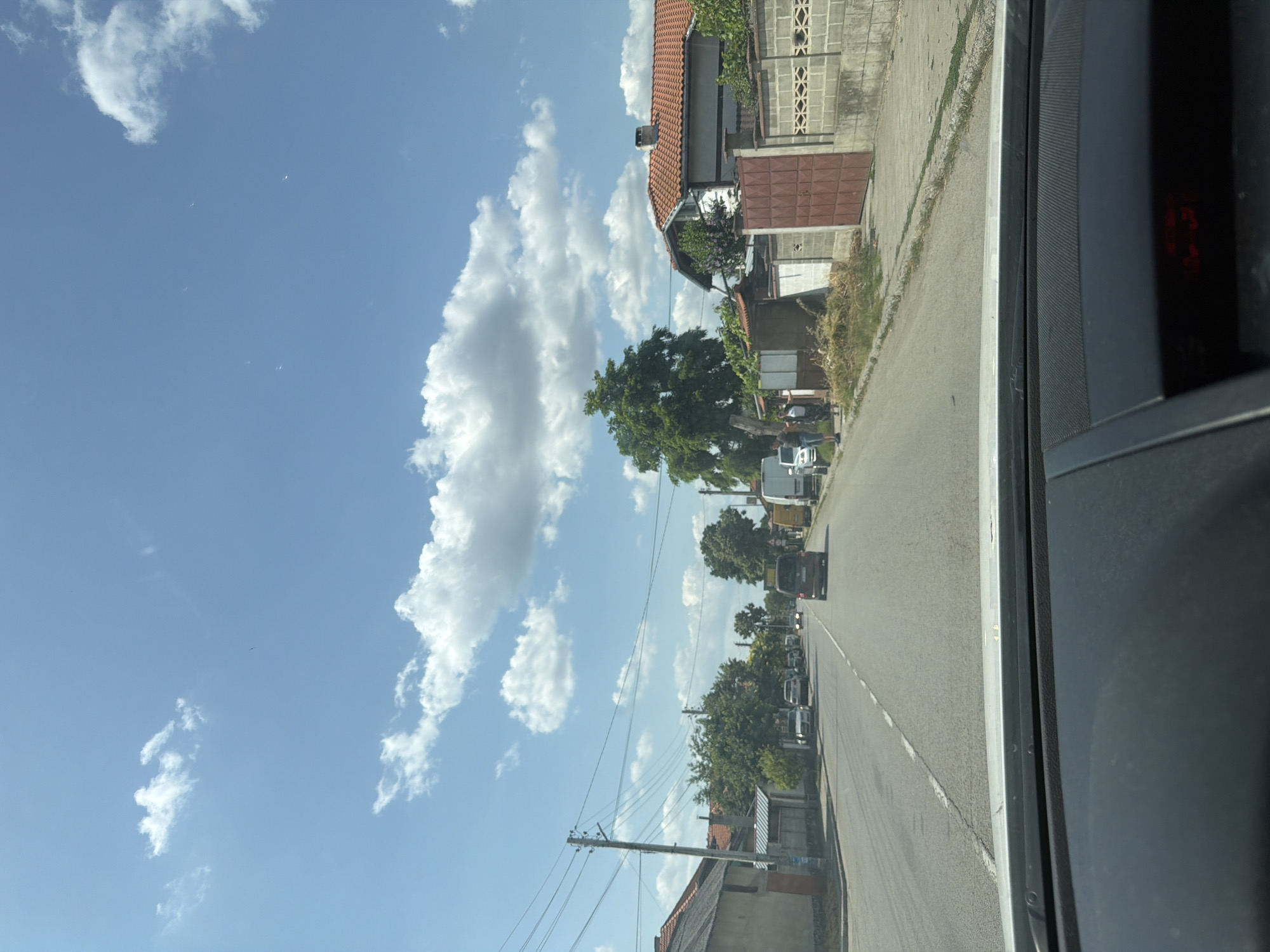 The image depicts a street scene in Tsar Kaloyan, Bulgaria, on a sunny day with scattered white clouds. The view is from inside a vehicle, looking forward through the windshield at the road ahead. The asphalt road is flanked by buildings and vegetation. On the left side of the road, there are houses with tiled roofs and a power pole with numerous wires. On the right side, there are more houses, some with brick walls and red-tiled roofs, and a large green tree. Several vehicles are visible on the road, including cars, vans, and a bus. Some people are standing on the sidewalk on the right side of the road, near a gate. The overall impression is of a residential area on a clear day.