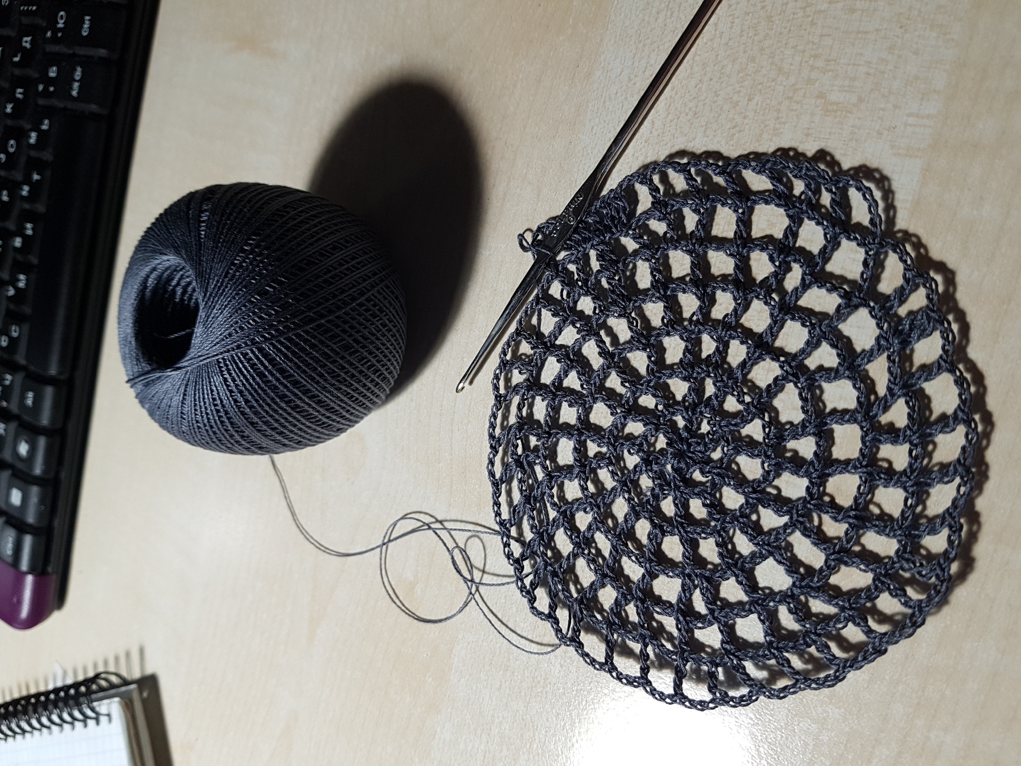 A high-angle shot captures a crafting scene on a light-colored wooden surface. A ball of dark gray thread sits beside a half-finished circular crochet project of a similar color, with a metal crochet hook inserted in the work. To the left, a black computer keyboard is partially visible, and at the lower left corner, a small notebook with a black spiral binding lies open. The scene is bathed in natural light, casting shadows that suggest the image was taken indoors, near a window. There is no evidence to identify the image's location as Saint Petersburg, Russia.