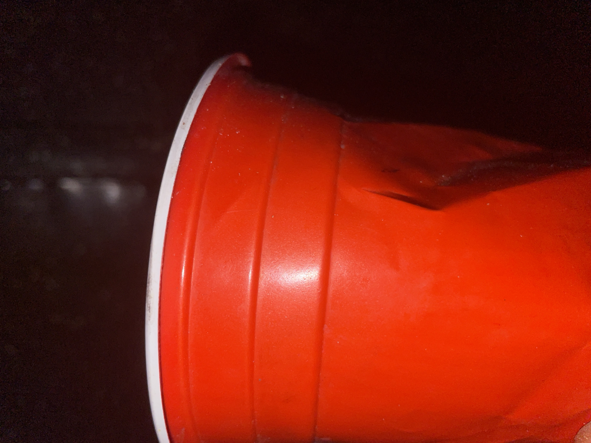A close-up, waist-up, interior shot shows a red plastic cup with a white rim against a dark, blurred background. The cup is slightly crushed and appears to be made of thin plastic. The surface of the cup has horizontal ridges and some white specks, possibly dust or residue. The lighting is dim, creating shadows and highlighting the texture of the cup. The location is not specified in the image.