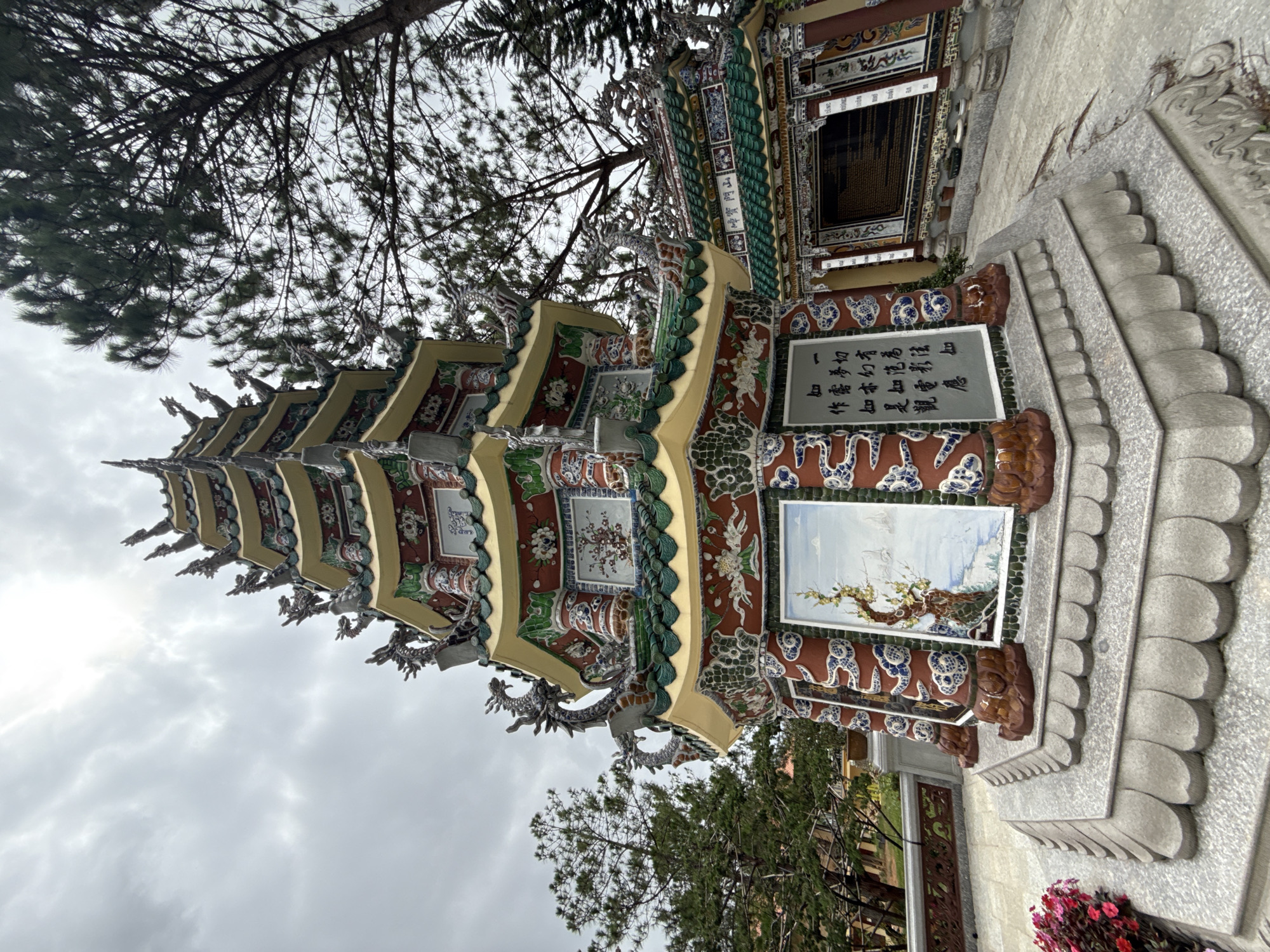 A low-angle, wide shot captures a richly decorated, multi-tiered pagoda with intricate dragon motifs and colorful ceramic tiles. The pagoda's roof is adorned with green tiles and decorative figures. Beneath the pagoda's multiple levels, artistic panels depicting landscapes and calligraphy are visible. The structure stands on a stone pedestal with layered, scale-like decorative elements. In the foreground, a cluster of pink flowers adds a splash of color to the scene. To the right of the pagoda, another building with a similar green tiled roof and decorative elements can be seen, featuring a large black plaque with text. The background is dominated by the canopy of tall pine trees and a cloudy sky. The overall setting appears to be a tranquil temple or spiritual site, likely in Dalat, Vietnam, known for its Buddhist temples and pagodas. There are no people visible in the image.