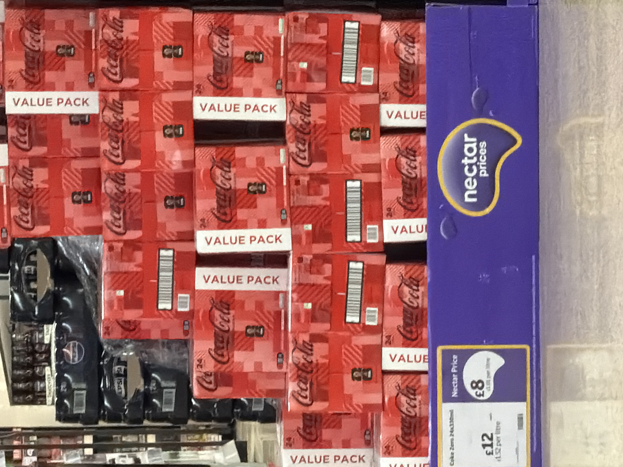 This image depicts a supermarket shelf stocked with Coca-Cola "Value Pack" 24-can cartons. The cartons are red with white "VALUE PACK" lettering and Coca-Cola branding. To the right of the Coca-Cola cartons is a blue "Nectar Prices" sign advertising "Coke Zero 24x330ml" for £8 (£1.52 per litre) as a Nectar Price with a standard price of £12. To the left, partly visible, are other products on the shelf, including some black packaged goods. Based on the "Nectar Prices" logo, the location is likely a Sainsbury's supermarket in Pity Me, United Kingdom, though this can't be conclusively determined from the image alone.