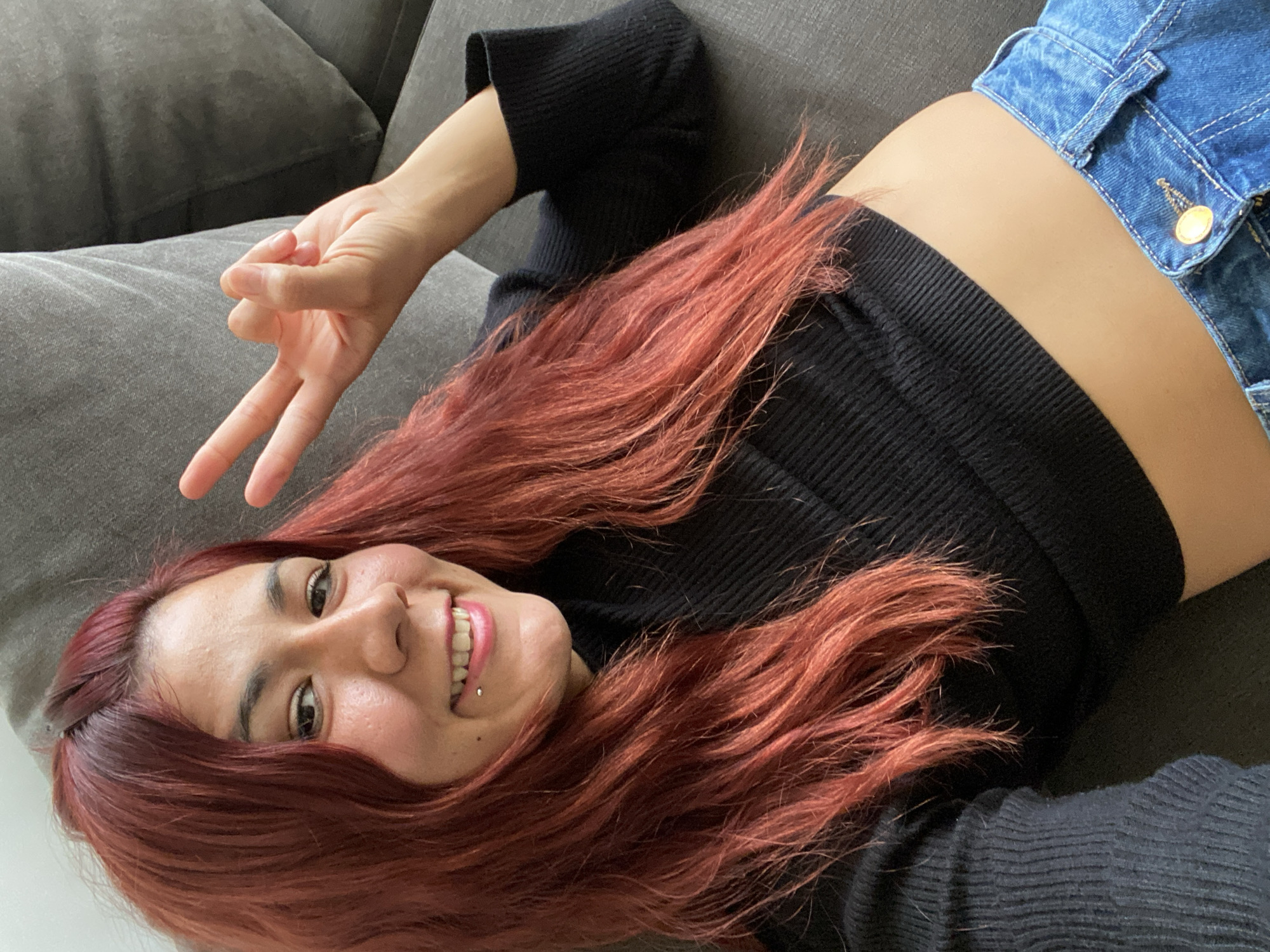 A woman with auburn hair is lying on a gray couch, smiling and holding up a peace sign. She is wearing a black long-sleeved crop top and denim shorts. A lip piercing is visible. The location appears to be indoors, with a gray couch and a gray pillow in the background. The specific location in Buenos Aires, Argentina, cannot be determined from the image.