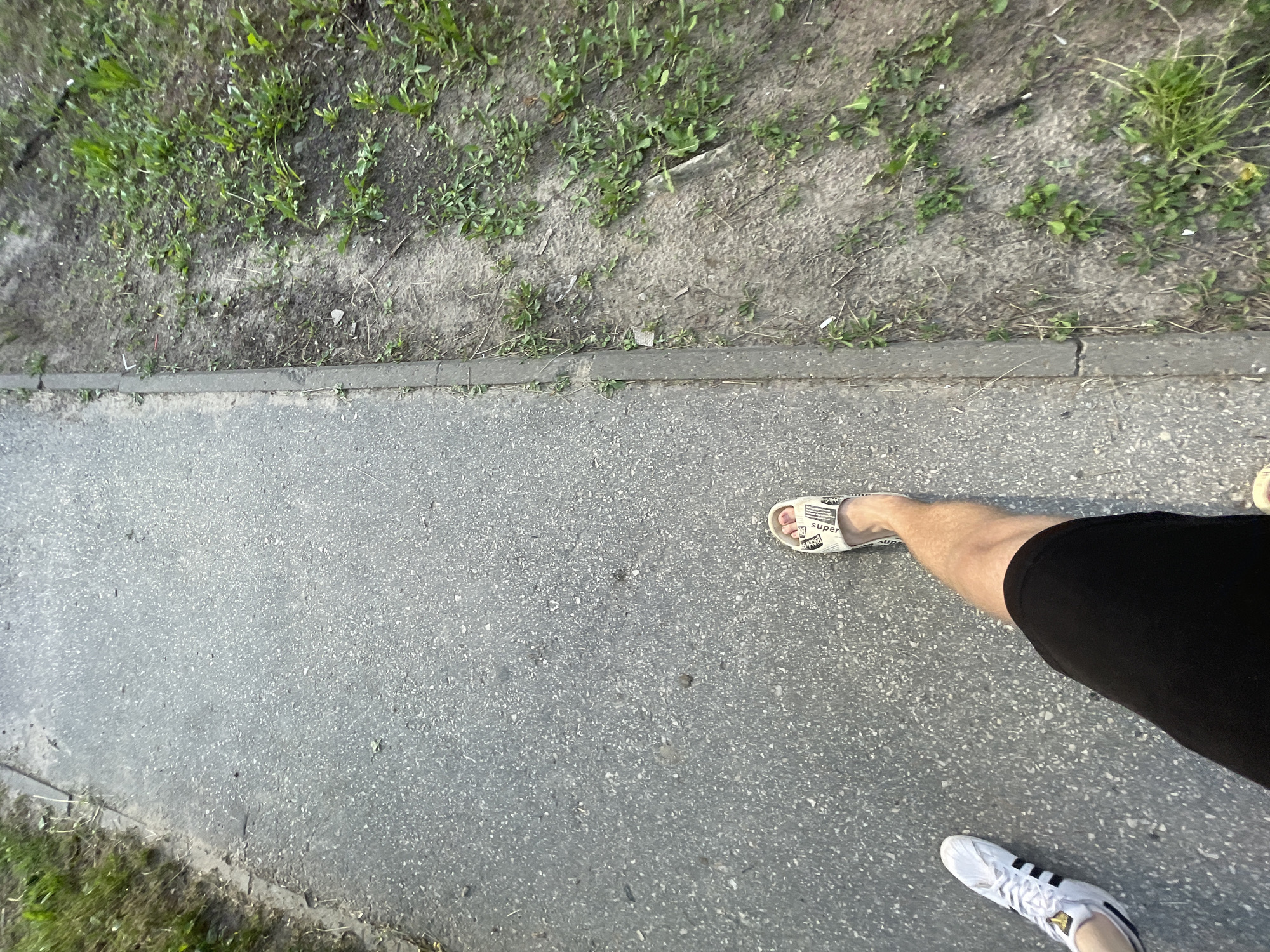 In Красные Баки, Russia, a person walks on a paved path next to a grassy area. The path is made of asphalt with visible aggregate. To the right of the path is a dirt area with sparse green vegetation and some dry grass. The person is wearing black shorts and two different types of footwear: a white Adidas sneaker on their left foot and a white slide sandal on their right foot. The image is taken from a first-person perspective, looking down at the ground and the person's legs and feet.