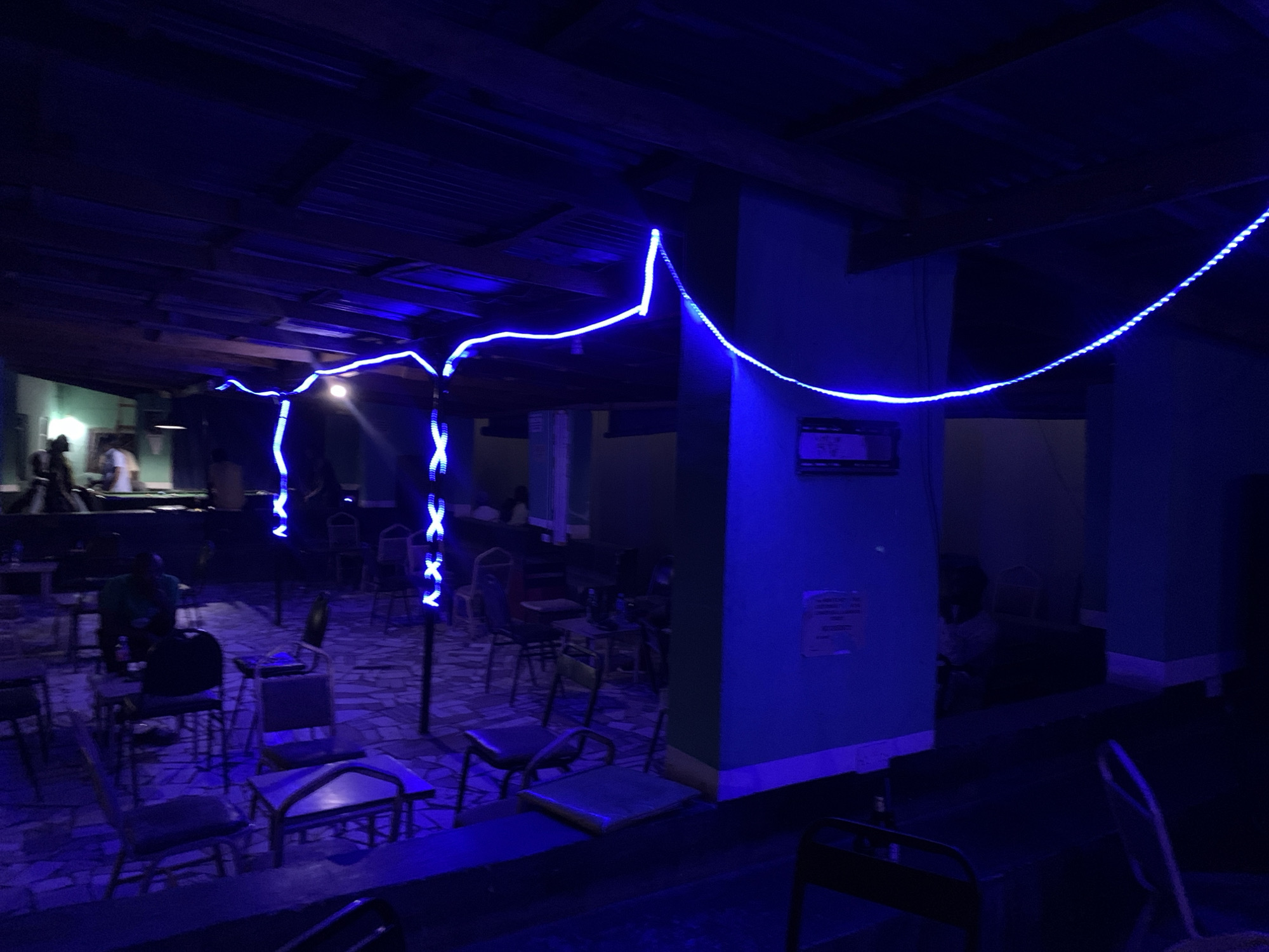The image shows an interior space, possibly a bar or lounge, in New Stadium, Nigeria. The room is lit with blue neon lights strung across the ceiling. There are several tables and chairs arranged throughout the space, and a few people are seated at some of them. A raised platform is visible in the background, where people are standing or walking. The overall atmosphere is dark and dimly lit.