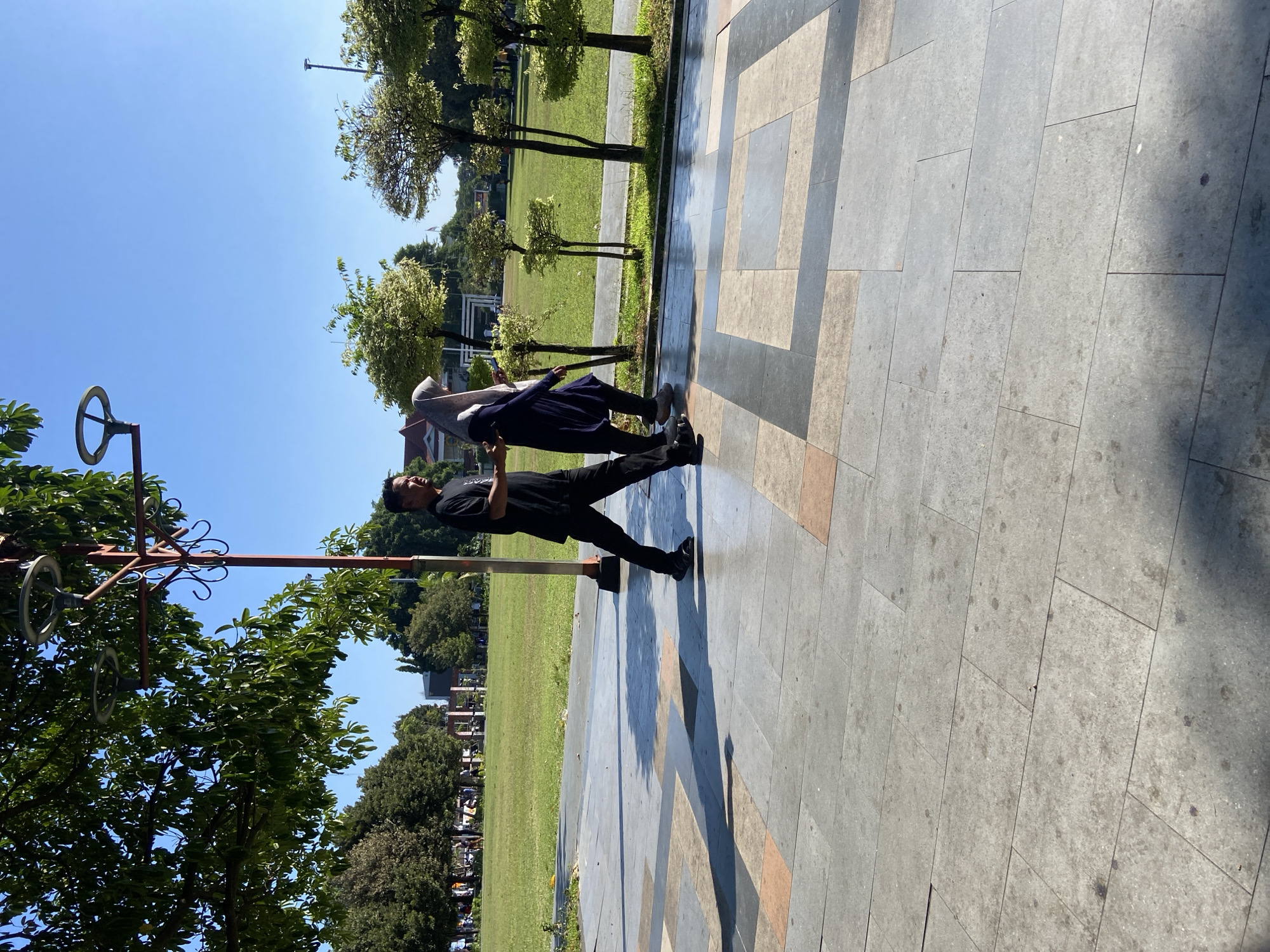A man and a woman are walking on a paved walkway in a park. The man is wearing a black t-shirt and black pants, and the woman is wearing a dark blue dress and a light gray hijab. They are walking side-by-side and appear to be engaged in conversation. In the background, there is a large grassy field with trees and a few buildings. The sky is clear and blue. The walkway is made of large, square tiles in various shades of gray, beige, and orange. The man is casting a long shadow to his left. The overall scene suggests a pleasant day in a public park in Kecamatan Pati, Indonesia.