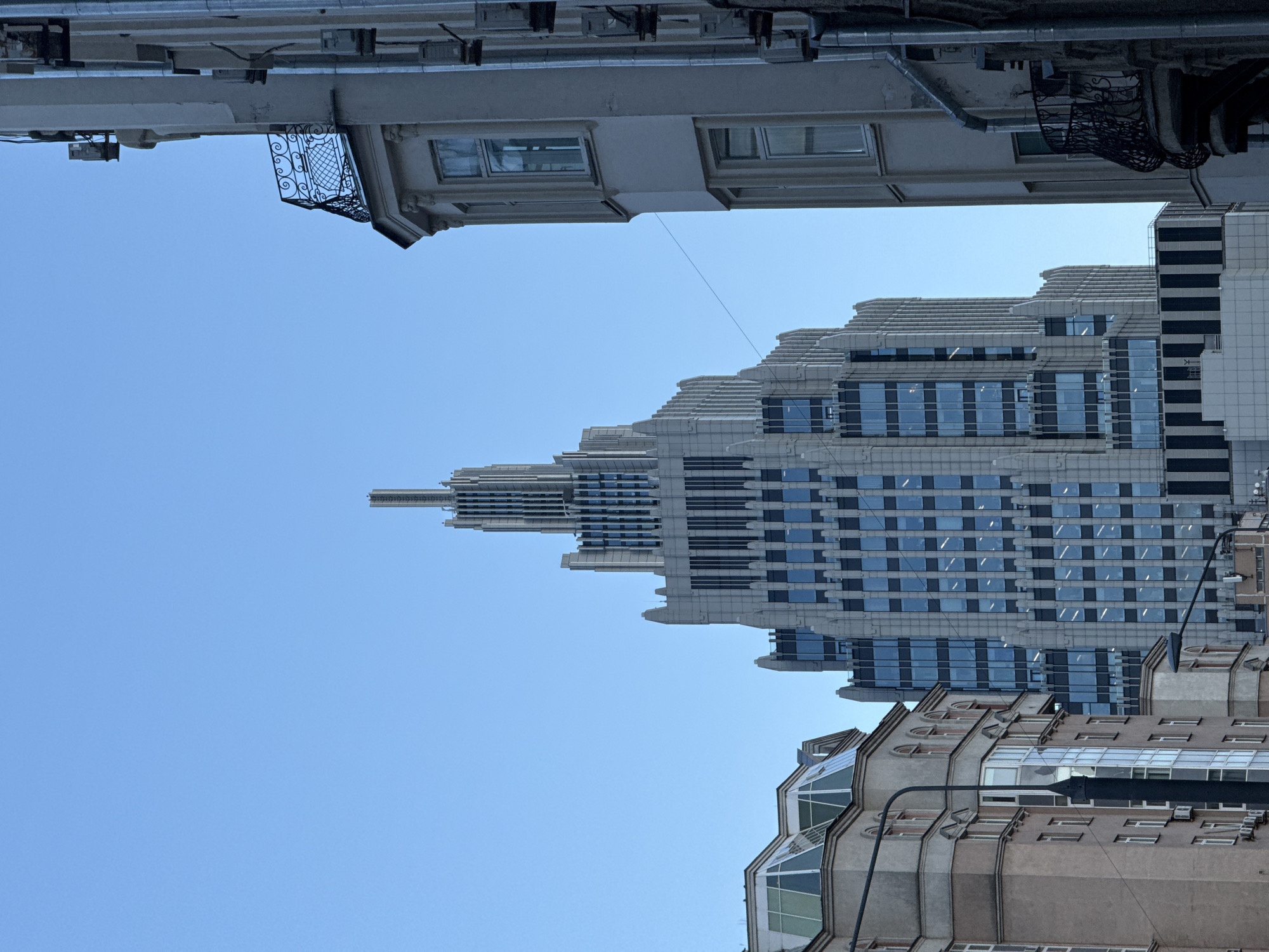The image presents an upward-looking view of an urban cityscape in Moscow, Russia, dominated by architectural structures set against a clear, bright blue sky.
In the center-right, a prominent modern skyscraper rises, characterized by a striking stepped or terraced design. Its facade features a repeating pattern of rectangular windows, many reflecting the sky, interspersed with light-toned horizontal bands and panels. The building tapers gracefully towards its upper sections, creating a distinctive profile.
To the bottom of the frame, a segment of an older building is visible, showcasing a more classic architectural style with arched windows and decorative elements, including sections with large, multi-paned glass. From the upper-left, another traditional building partially enters the frame, featuring an ornate wrought-iron balcony and visible utility pipes or structural components beneath its overhang.
The scene is illuminated by natural light, indicating daytime with perfectly clear weather, as evidenced by the unblemished, uniform blue sky. There are no people visible, and no specific activities are taking place, placing the focus squarely on the diverse urban architecture. No text is discernible in the image.
