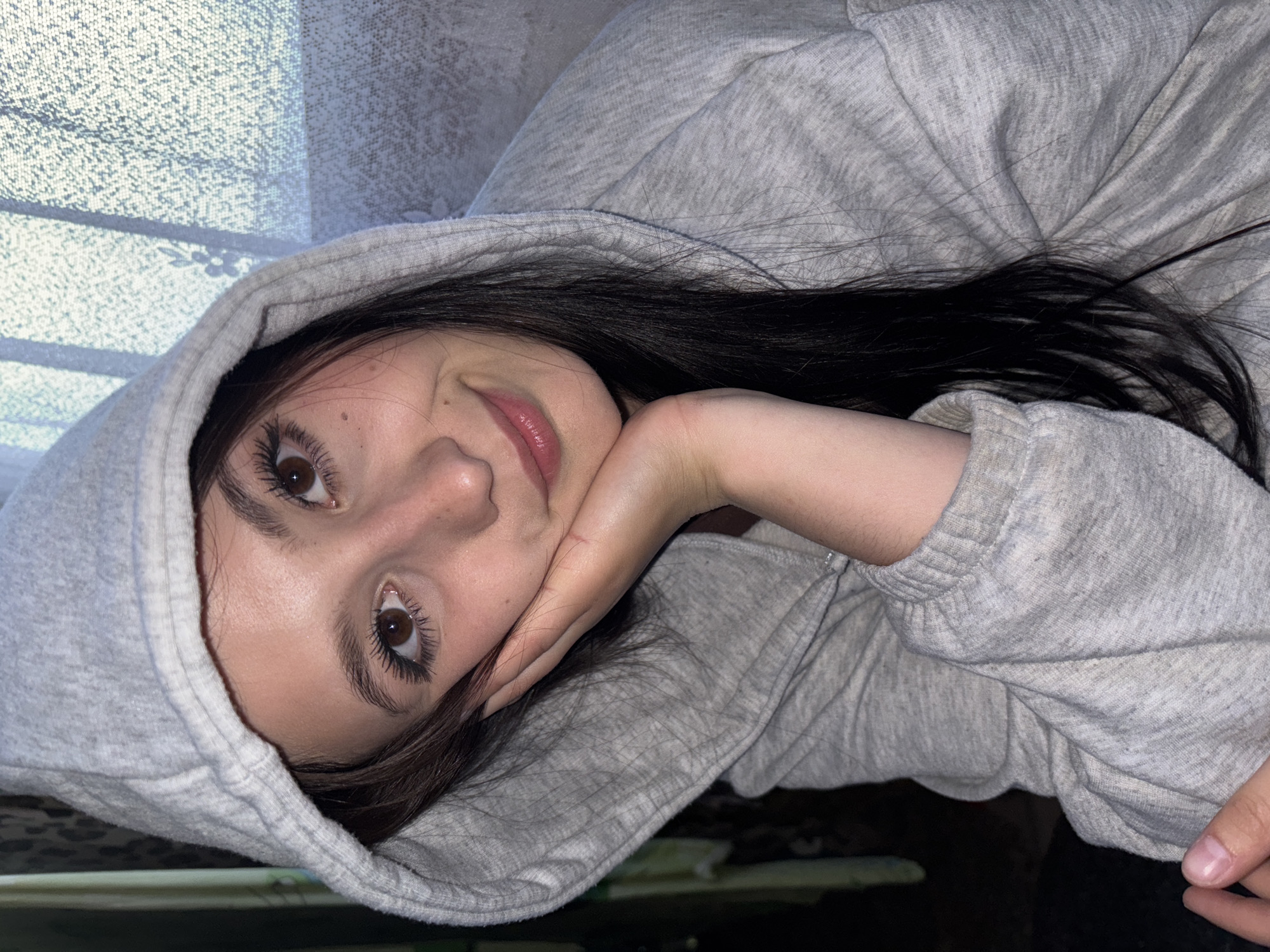 This close-up portrait features a young individual, likely female, with long dark hair and brown eyes, looking directly at the camera with a soft smile. She is wearing a light grey hooded sweatshirt, with the hood pulled up and framing her face. Her head is tilted, and her left hand rests gently against her cheek, supporting her chin.
The background suggests an indoor setting. To the upper left, natural light filters through a textured, light-colored fabric, possibly curtains or blinds, creating patterned shadows and highlights. This indicates it is daytime. The person's expression conveys a sense of ease and contentment. No text is visible in the image.