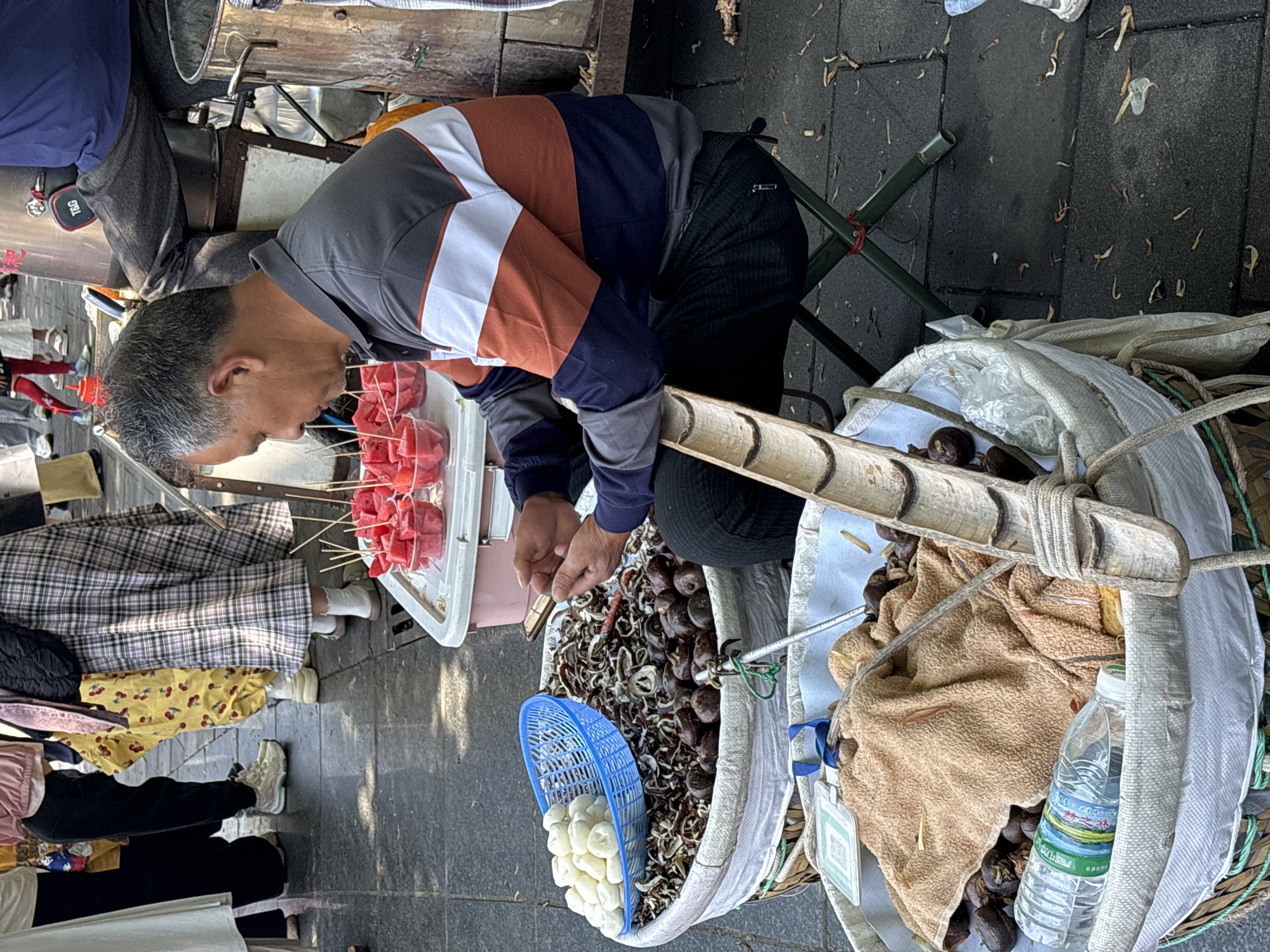 An elderly man sits on a green folding stool next to baskets filled with roasted chestnuts, peeled water chestnuts in a blue basket, and skewers of watermelon on a street in Jiulongpo District, China. He is wearing a multi-colored long-sleeved shirt and dark pants. Customers wearing various outfits stand nearby. A clear plastic water bottle sits in one of the baskets with the chestnuts.