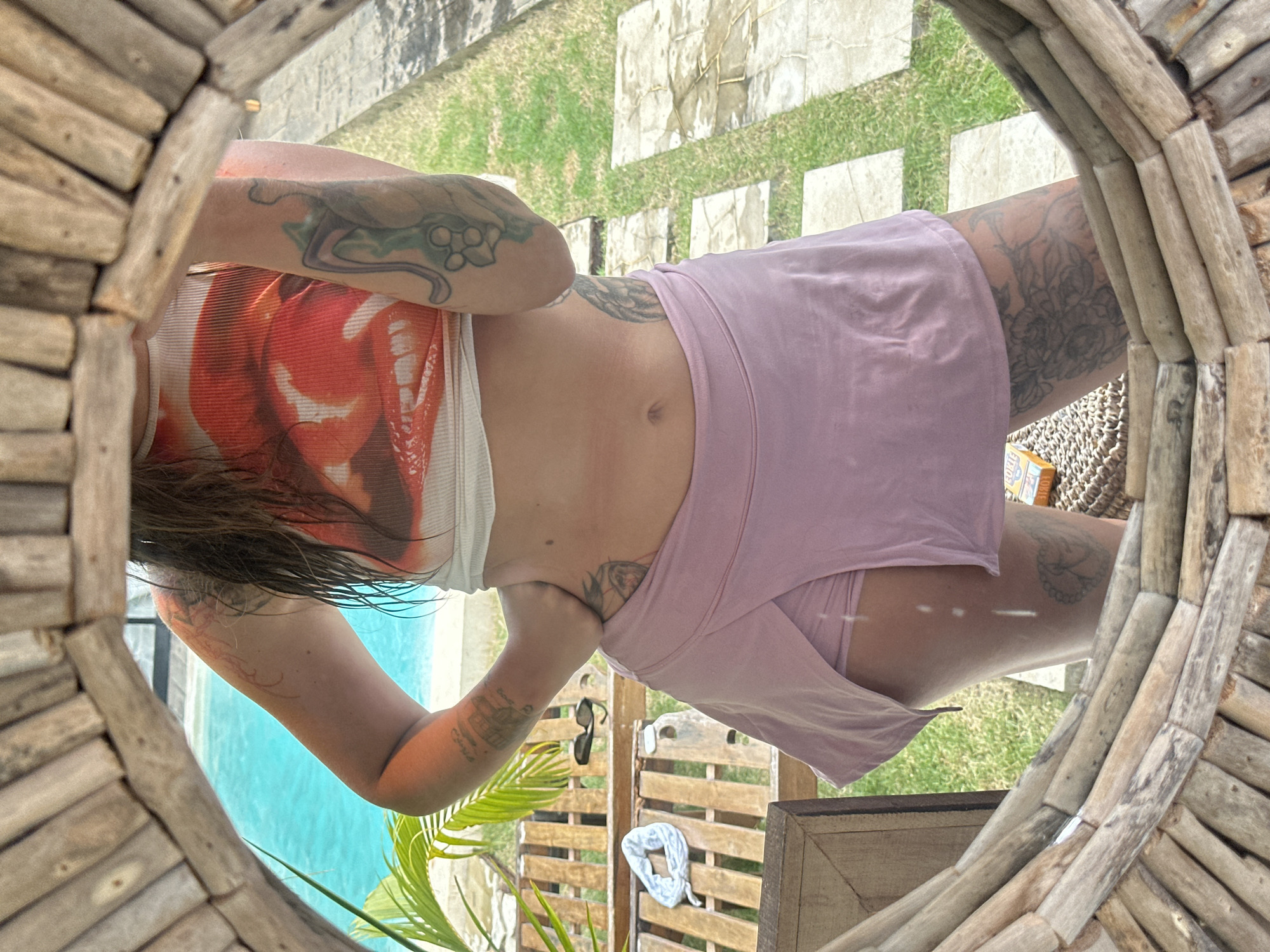 A person is reflected in a circular mirror with a wooden frame, showing their torso and upper legs. They are wearing a white ribbed tank top with a graphic print of red lips and a light pink skirt with a side slit. Tattoos are visible on their arms and legs. In the background, a swimming pool with bright blue water and lush green foliage can be seen, along with a wooden bench and stone pathway. The scene appears to be outdoors, possibly in a tropical or resort setting.