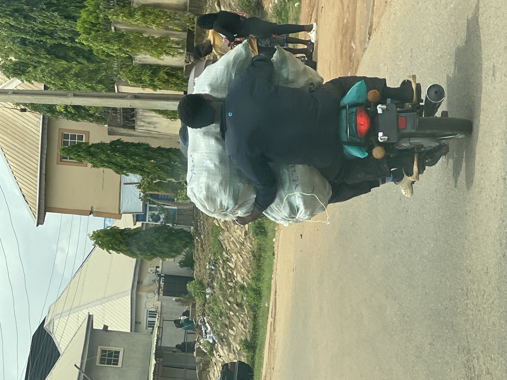 The image captures a street scene in Jos, Nigeria. Two individuals are on a motorcycle, with the passenger in the rear carrying a substantial load of large, white sacks strapped to their back and the motorcycle. The driver is wearing a dark blue shirt, and the passenger is also dressed in dark clothing. Behind them, another man in a yellow shirt and a woman in black leggings and a black top are visible, standing near a pile of debris and overgrown vegetation. The background features several residential buildings with distinctive pitched roofs and walls, interspersed with tall, slender trees. A power pole stands to the right of the motorcycle. The road surface is asphalt, and the sky is overcast.
