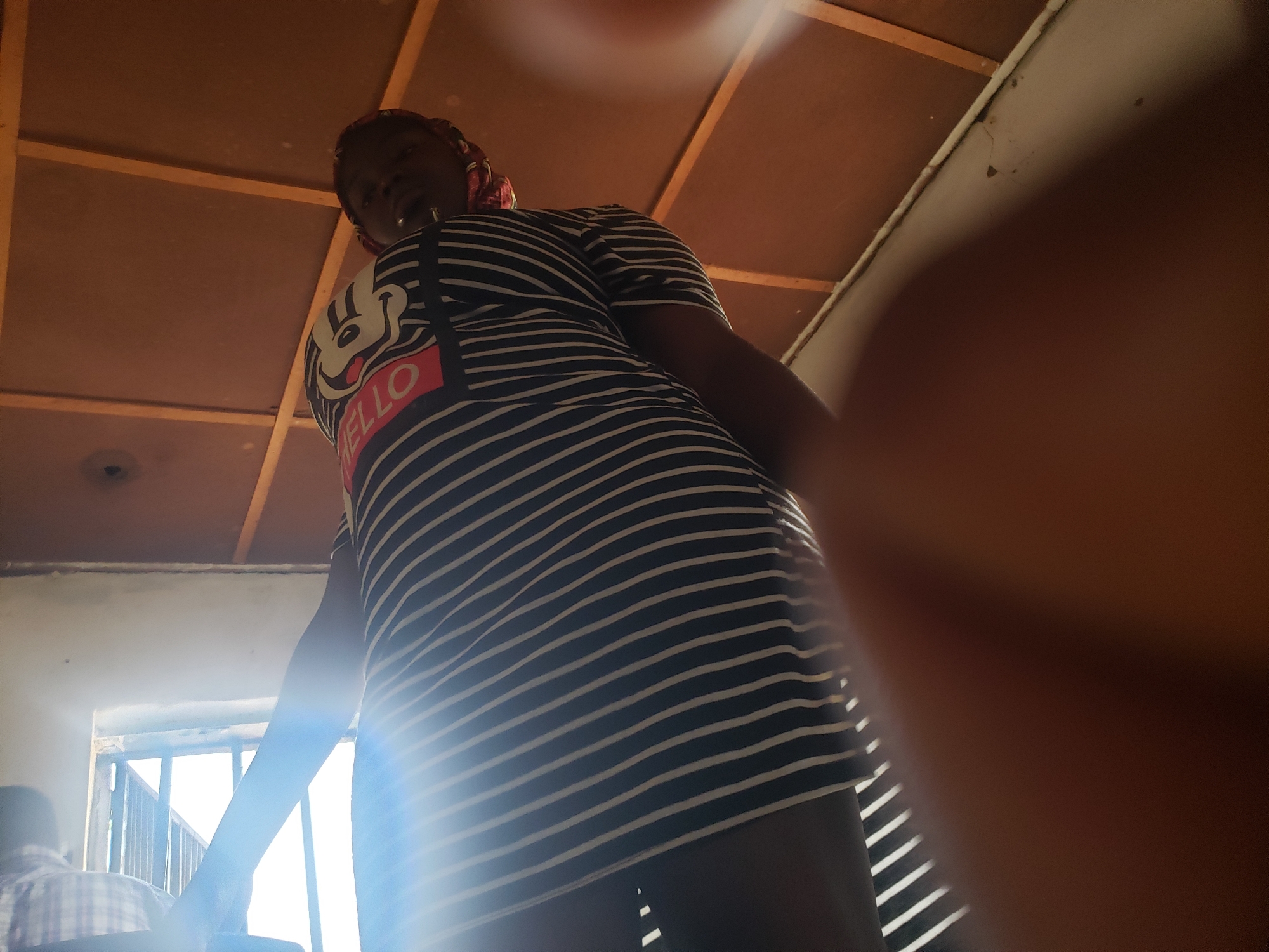 In Kachia, Nigeria, an individual, appearing to be a female, is captured from a low-angle perspective, looking upwards towards the camera. She is wearing a black and white striped dress with a graphic print on the chest featuring a cartoon character and the word "HELLO". Her head is adorned with a red headwrap. The background consists of a wooden ceiling with visible beams and a portion of a white wall. Sunlight streams in from the left, creating a lens flare effect and highlighting the stripes of her dress. To the left, a barred window is partially visible, with another person in a checkered shirt seated in front of it. The foreground is dominated by a blurred, reddish-brown object, likely a hand or finger, obscuring a portion of the image. The overall lighting is warm and diffused, with strong highlights from the sunlight.