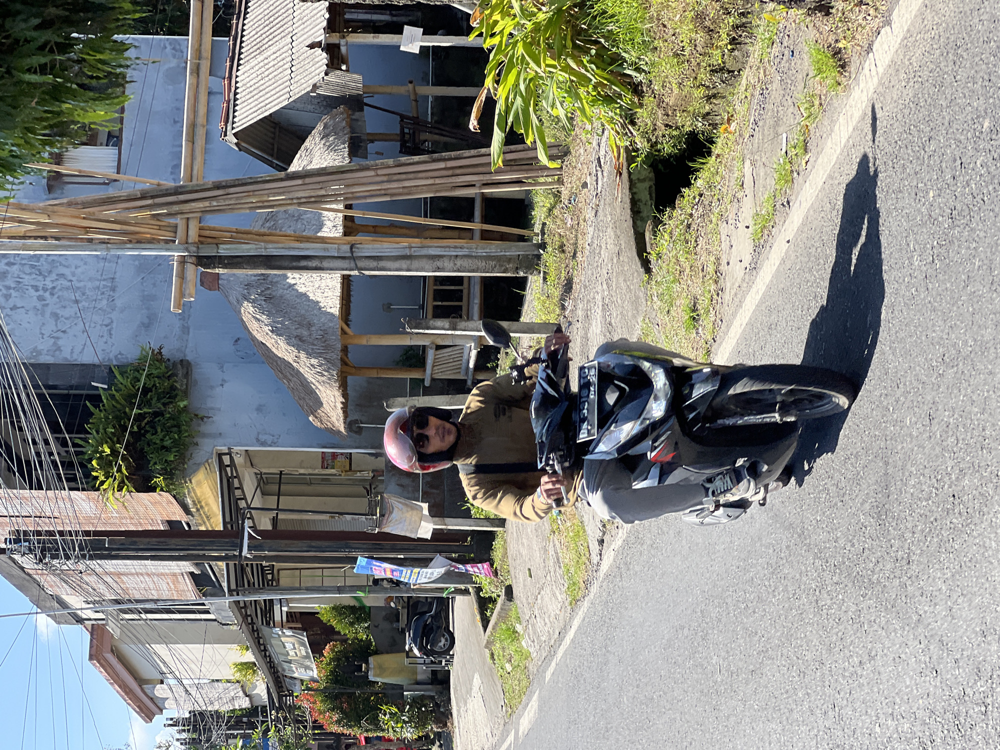 A man wearing a tan long-sleeved shirt, grey pants, a red helmet, and sunglasses rides a black and silver scooter on a paved road. He is looking forward, and his left hand is on the scooter's handlebars. The scooter has a license plate visible. In the background, there are buildings with balconies and awnings, electrical wires, bamboo structures, and thatched-roof huts. Greenery, including plants and trees, is present on the sides of the road and on the buildings. The scene appears to be in a tropical or subtropical urban environment. The location is the District of Ubud, Indonesia.