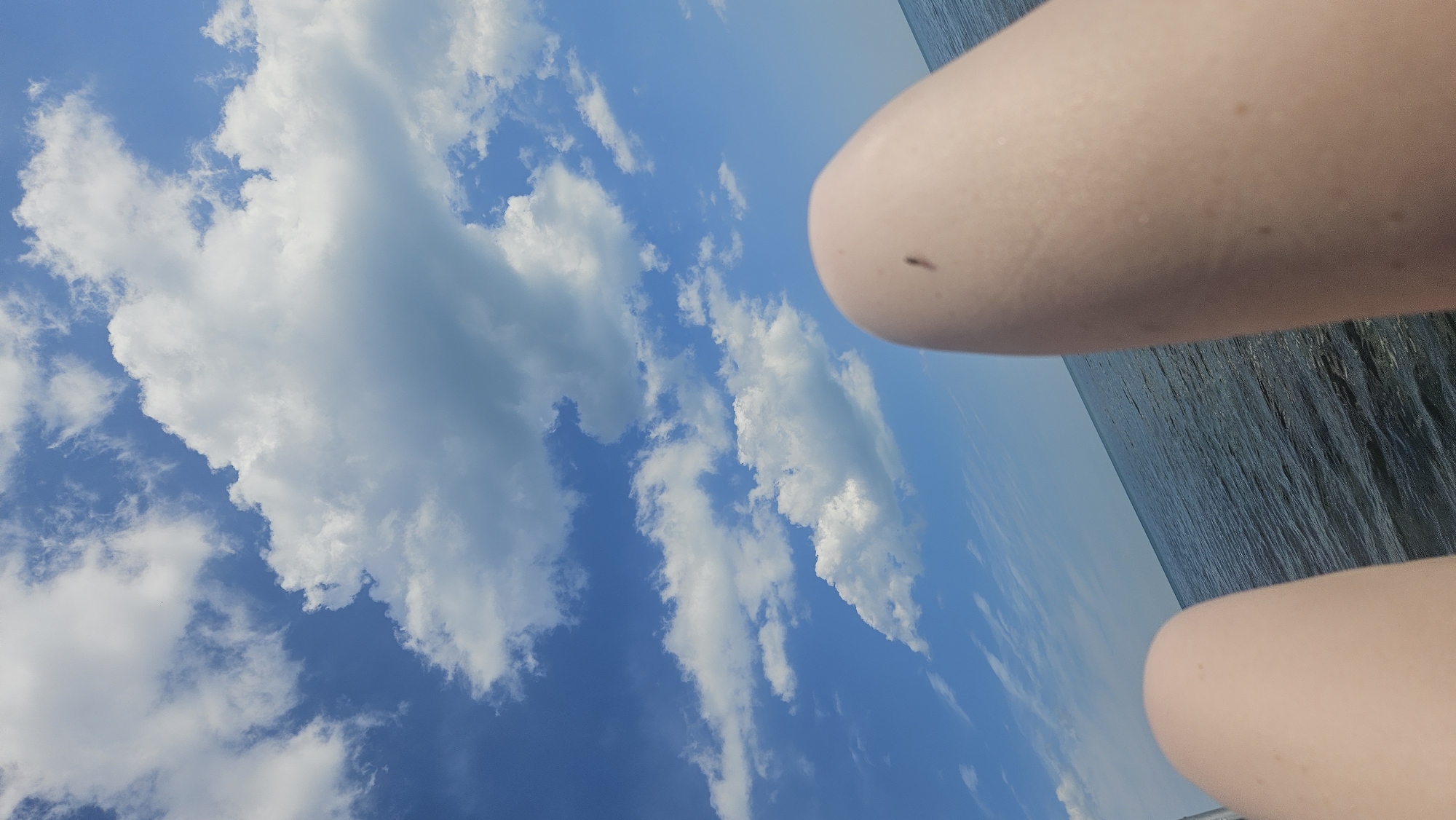 The image shows a close-up view of a person's legs against a backdrop of a blue sky with white clouds and a body of water. A small insect is visible on the skin of the right leg. The water appears to be a sea or a large lake, with gentle waves. The perspective suggests the person is looking up at the sky or taking a selfie from a low angle. No specific location in the city of Dyurso, Russia can be determined from the image.