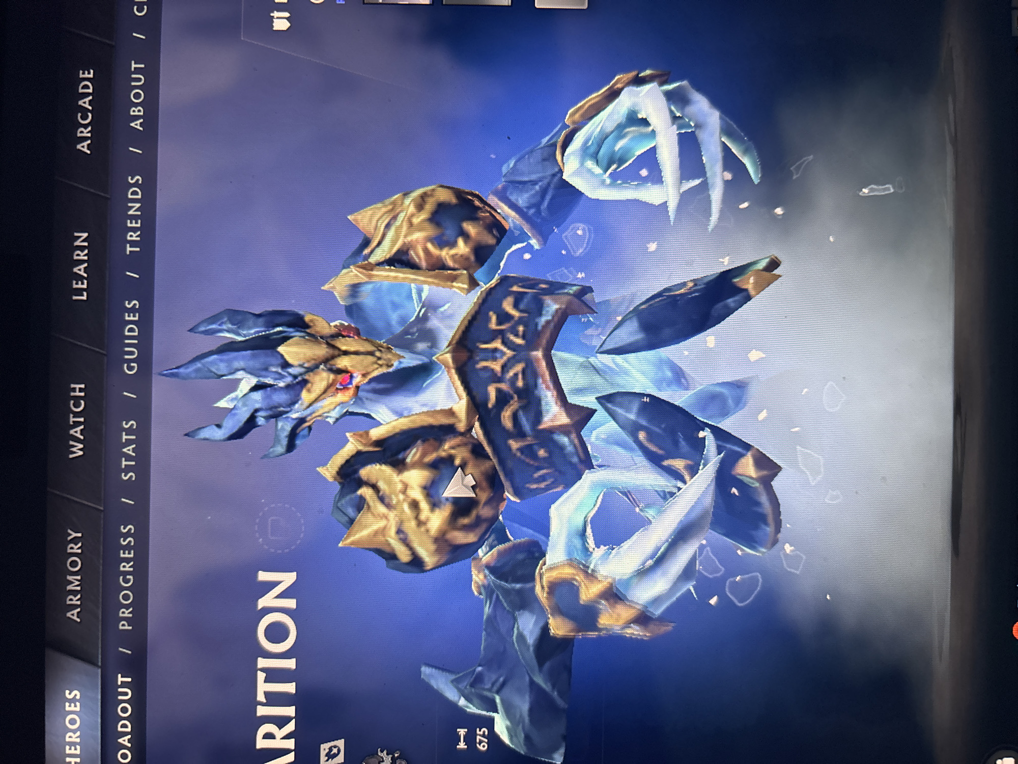 An image, captured at an angle, displays a digital screen or monitor featuring what appears to be a character or creature from a video game. The main subject is a stylized, fantasy-like entity with a dominant color scheme of deep blue, gold, and light blue/white glowing accents. It features sharp, claw-like appendages that emit a frosty or ethereal glow, and its body is adorned with intricate golden armor-like plating. The character's head, visible on the left, has a reddish-gold eye and spiky blue features.
The background is a dark blue, gradient-like digital interface, with subtle smoky or misty effects and small, glowing particulate matter around the character. The image is taken in portrait mode but appears rotated, showing the screen's content horizontally.
Along the left side of the screen (which is the top of the image due to rotation), a vertical menu bar is visible with several options, including "HEROES", "LOADOUT", "ARMORY", "PROGRESS", "WATCH", "STATS", "LEARN", "GUIDES", "ARCADE", "TRENDS", "ABOUT", and a partially visible "C". A large word, "ARITION" (likely part of "APPARITION"), is displayed prominently in white text near the bottom center of the screen, indicating the character's name. Below this, a small numerical value "675" is visible. Some faint, partially visible text "ut" appears in the top right corner. The overall scene suggests a character display or selection screen within a gaming application.