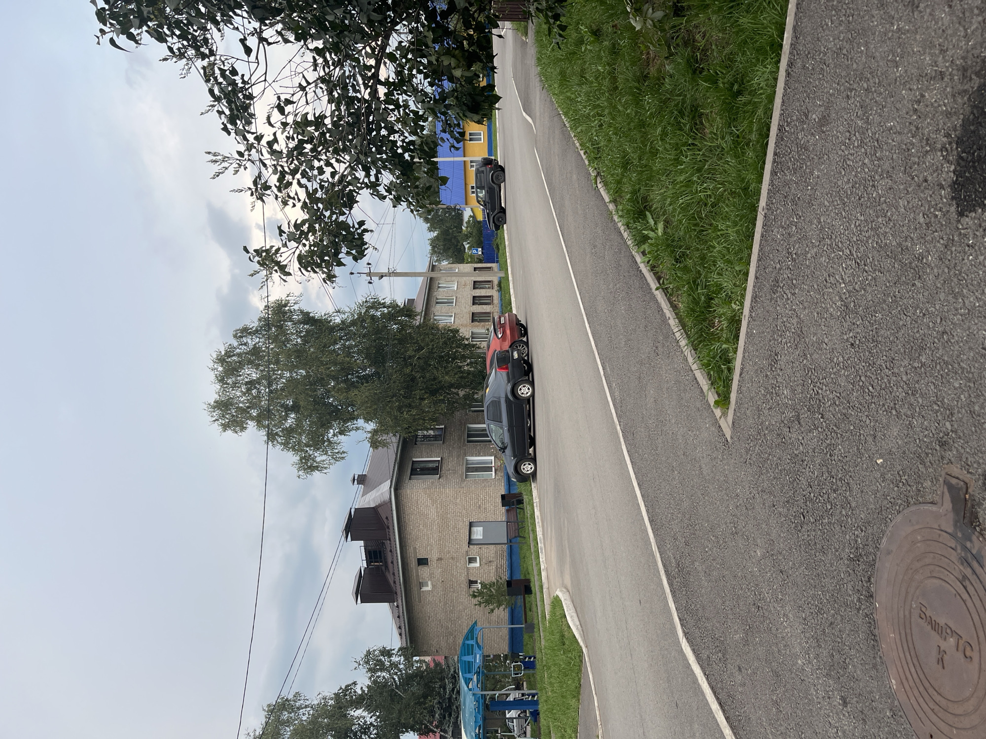 An exterior, eye-level, medium shot captures a residential street scene in Blagoveshchensk, Russia, under an overcast sky. The image is dominated by a two-story brick building with a dark, sloped roof. To the left, a blue awning covers a small waiting area with seating. Several cars are parked along the road, including a dark sedan, a red sedan, and a black SUV. A bright yellow house with a blue roof is visible in the distance to the right. Lush green grass lines the side of the road and surrounds the buildings. A manhole cover is visible in the foreground on the asphalt. The overall impression is one of a quiet, suburban street.