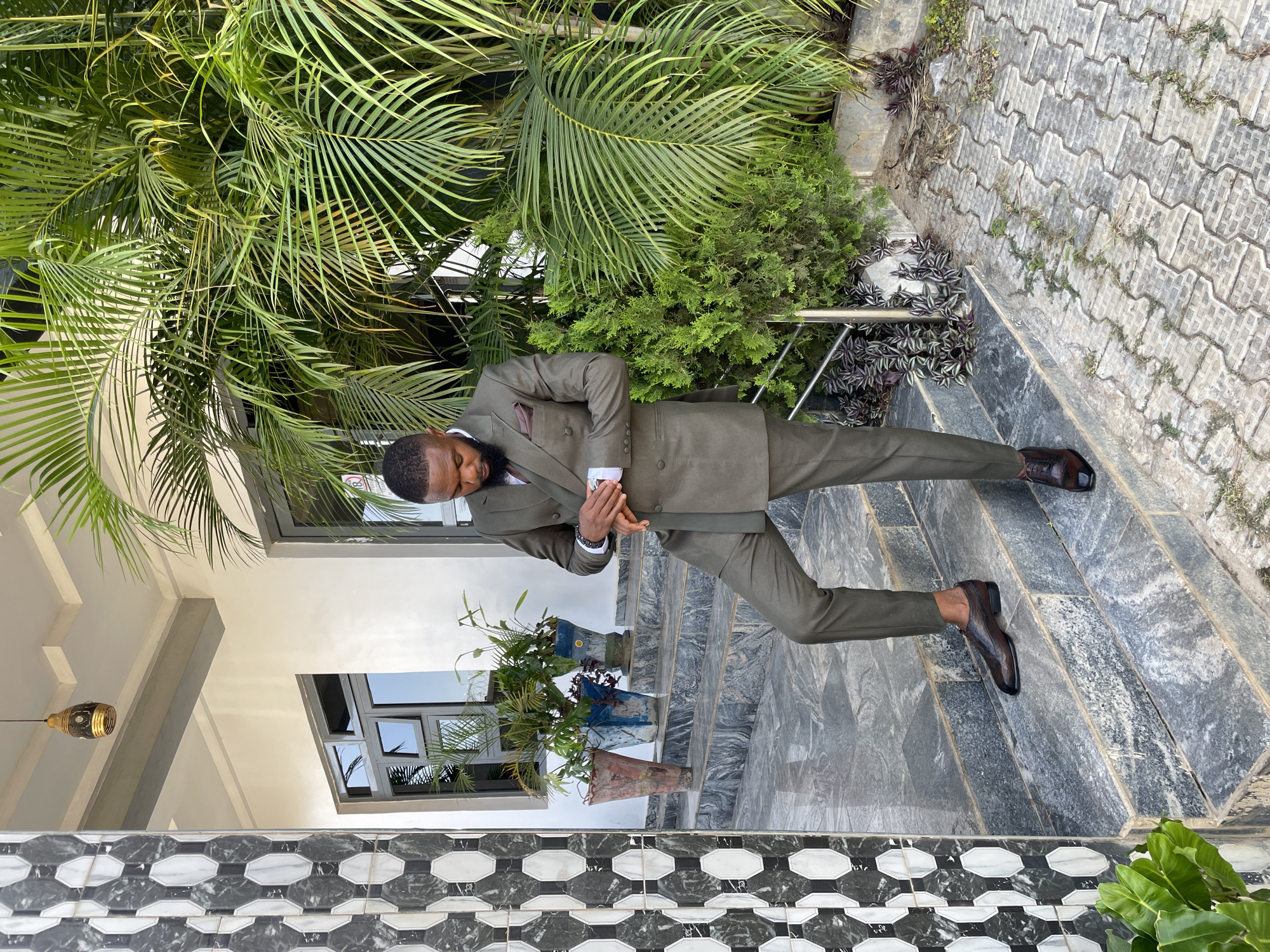 A man in a well-tailored, olive green suit is the central subject of the image. He stands on a tiled outdoor staircase leading to a modern-style building with large windows and white walls. The tiled staircase is gray with a patterned border of alternating black and white polygons. Various plants frame the scene, including tropical foliage to the side and potted plants near the building's entrance. The man is looking down, holding something in his hands. The image might be taken in Jos, Nigeria, but that's not explicitly confirmed by elements in the image.