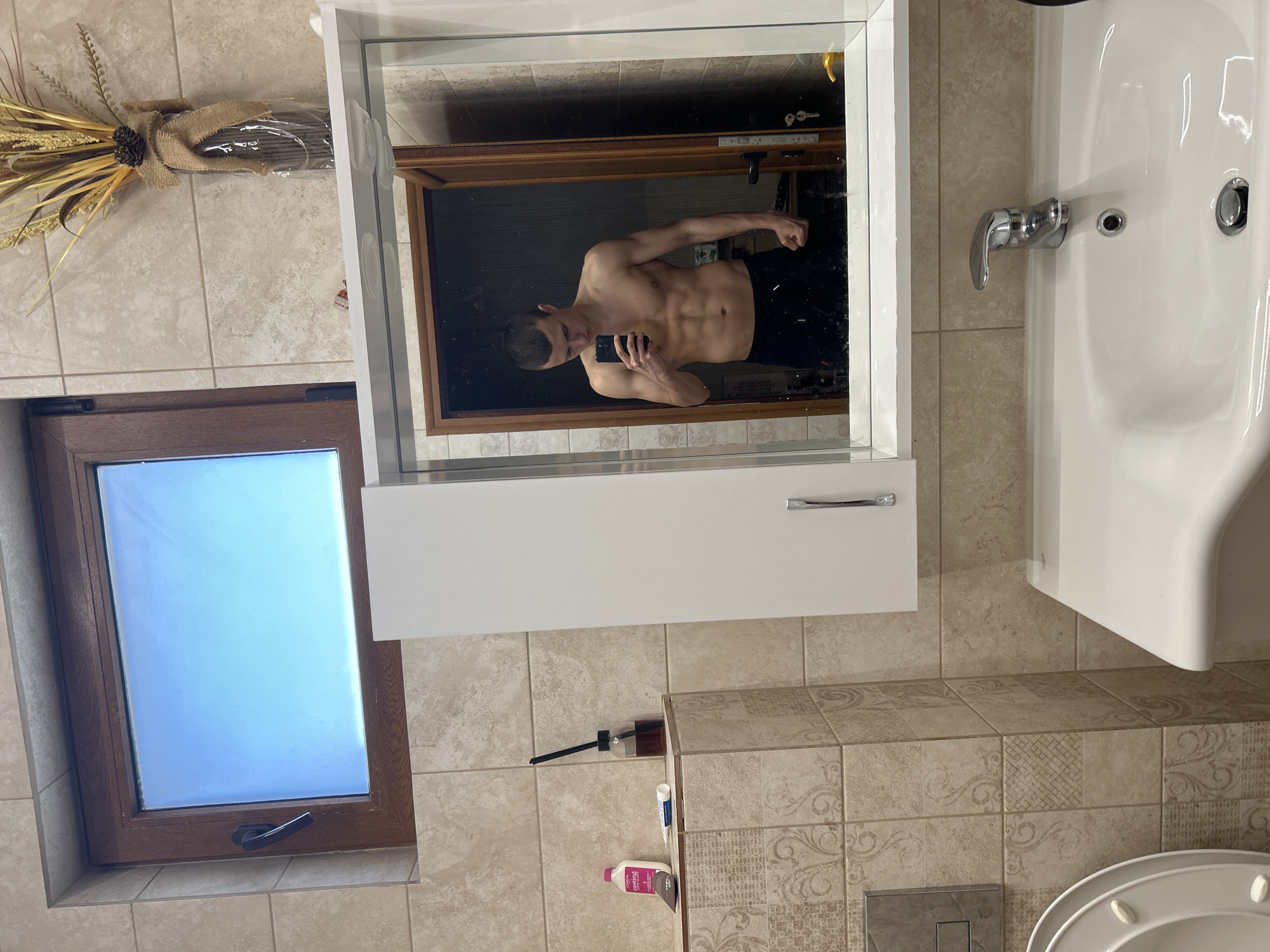 The image shows a bathroom in an unknown location, potentially Burgas, Bulgaria. A man is visible in the mirror, shirtless, taking a photo of himself with a phone. He is wearing black shorts. The bathroom has a sink, a toilet, and a window. There is a decorative wheat bundle on the wall, and various toiletries are on the floor. The walls and floor are tiled in a light beige color. The window is small and rectangular, and appears to be letting in daylight.