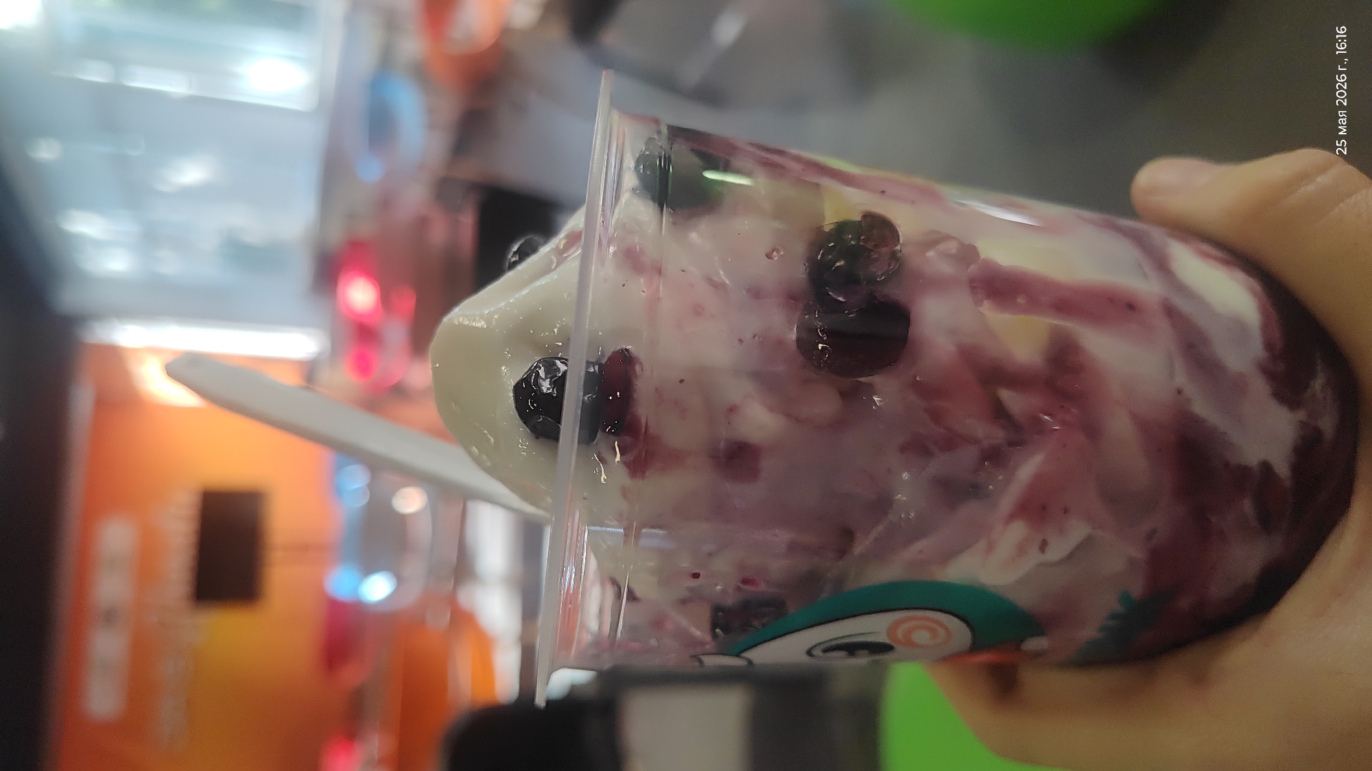 The image shows a close-up of a plastic cup containing what appears to be a yogurt parfait with blueberries and purple swirls, held by a hand. A white plastic spoon is inserted into the parfait. The background is blurry but suggests an indoor environment with orange, blue, and white fixtures. The date and time "25 мая 2026 г., 16:16" (May 25, 2026, 4:16 PM) are visible in the upper right corner. There is no information to infer this image's location in Almaty, Kazakhstan.