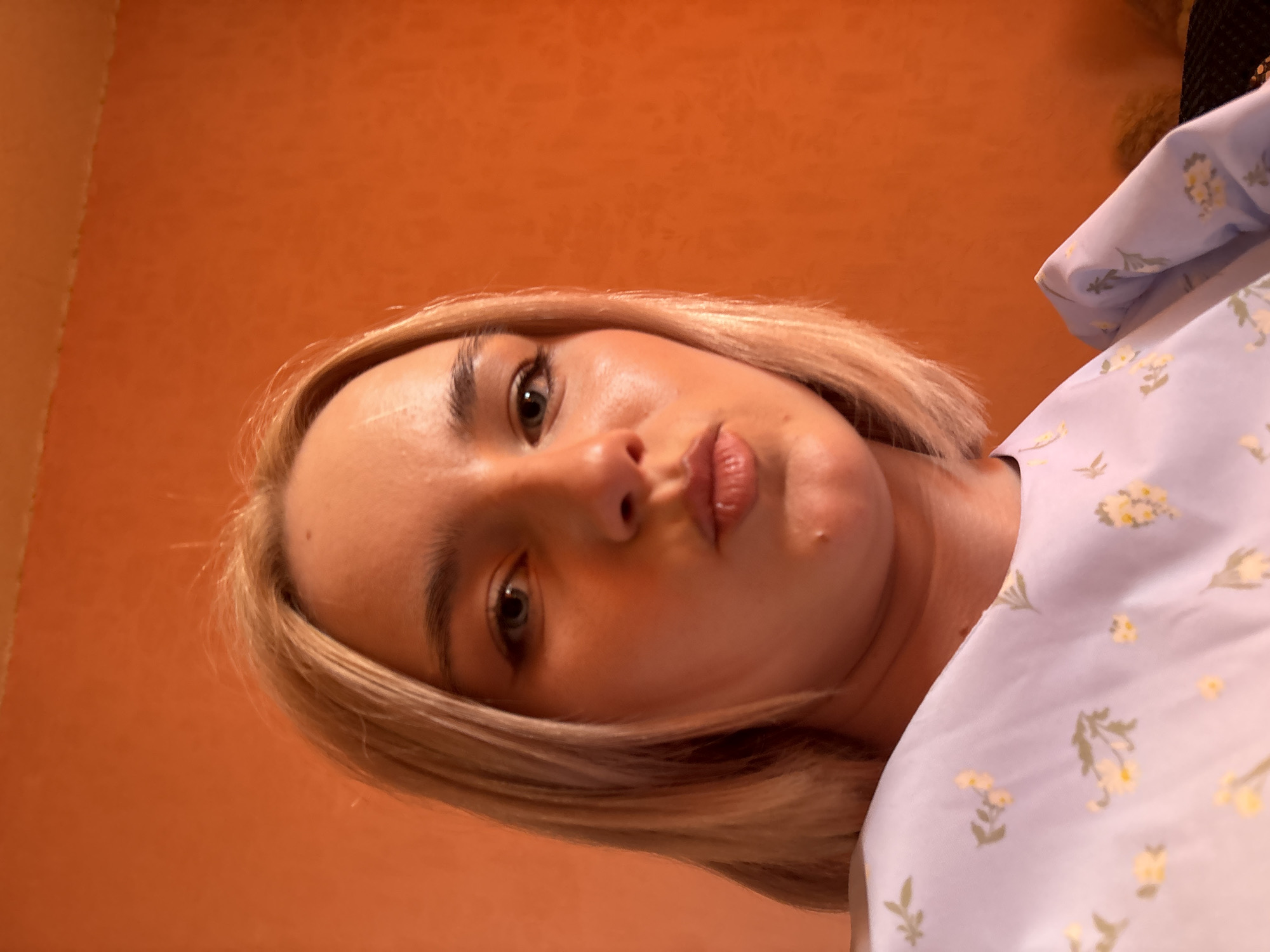 A close-up, low-angle self-portrait features a young woman with blonde, chin-length hair. She is looking directly at the camera with dark eyes, her lips slightly parted in a pout. Her face is well-lit, and her skin appears fair, with defined eyebrows. She is wearing a light-colored top or dress, possibly lavender or light blue, adorned with a delicate pattern of small yellow and green floral elements. The background consists of a solid, warm orange or terracotta-colored wall, which is largely out of focus, creating a shallow depth of field that emphasizes the subject. The lighting is bright and even, indicating an indoor setting. No text is visible in the image.