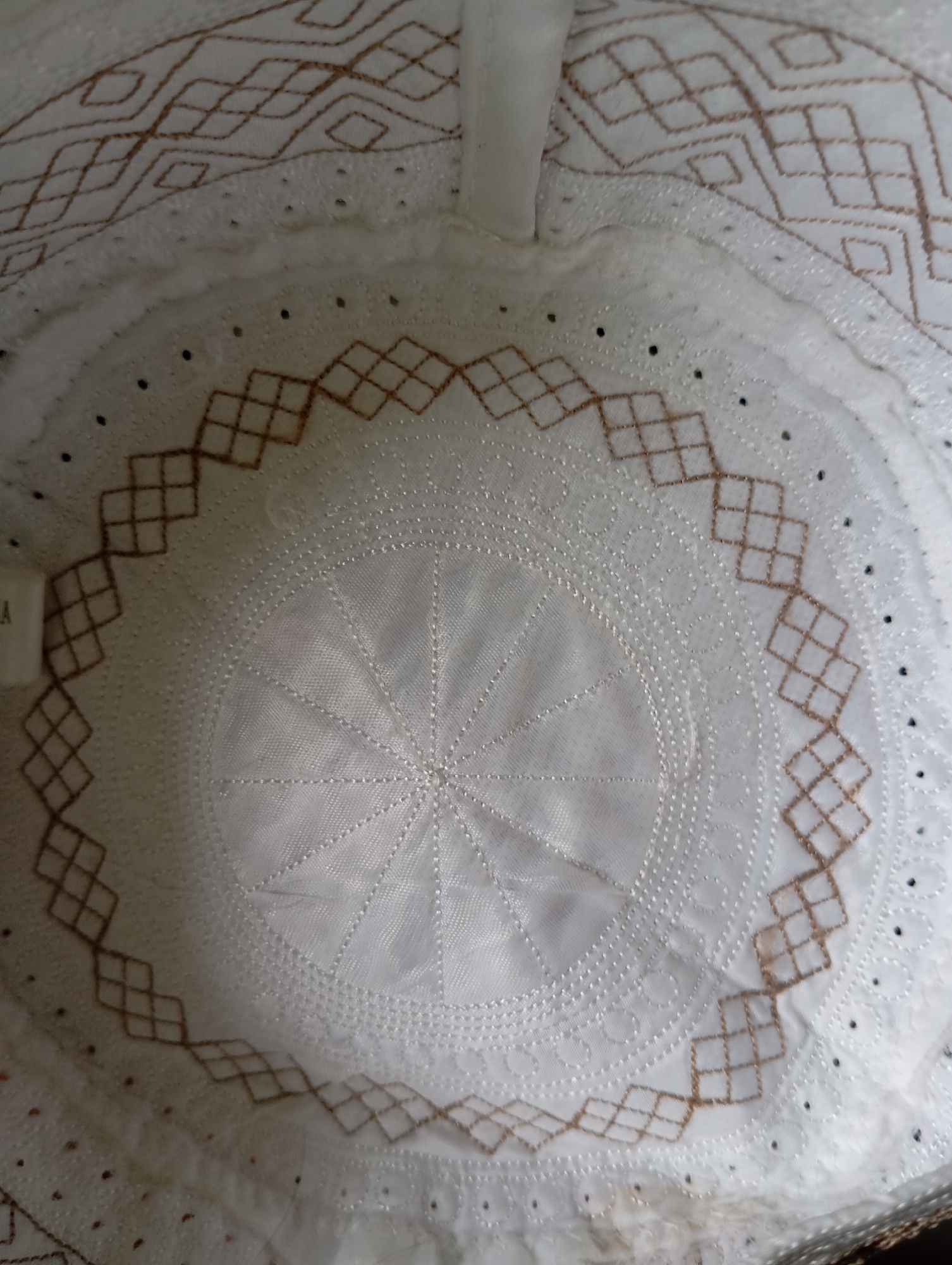 A close-up, top-down shot shows the interior of a white cap with intricate brown embroidery. The embroidery features geometric patterns, including diamonds and repeating lines, arranged in concentric circles around a central starburst pattern. The fabric appears to be cotton or a similar breathable material, with small perforations visible in some areas. A small white tag with faded black text is partially visible on the left side. The lighting is diffused, creating subtle shadows that highlight the textured embroidery. The background is blurred, suggesting the cap is resting on a surface.
