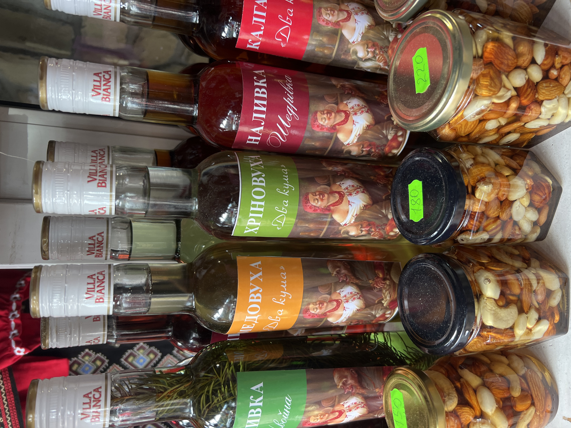 The image shows a market stall in Yaremche, Ukraine, displaying an assortment of homemade liquors in glass bottles and jars of nuts. The bottles are capped with white and gold seals, each bearing the "Villa Bianca" logo. Several bottles have colorful labels featuring illustrations of traditional Ukrainian figures and Cyrillic script. In the foreground, three glass jars are filled with various nuts, such as almonds and cashews, preserved in a liquid. Some jars have price tags attached to their lids, indicating "150," "180," and "220." The background reveals a glimpse of patterned textiles, suggesting a traditional Ukrainian setting within the market. The lighting appears to be natural, illuminating the products for sale.