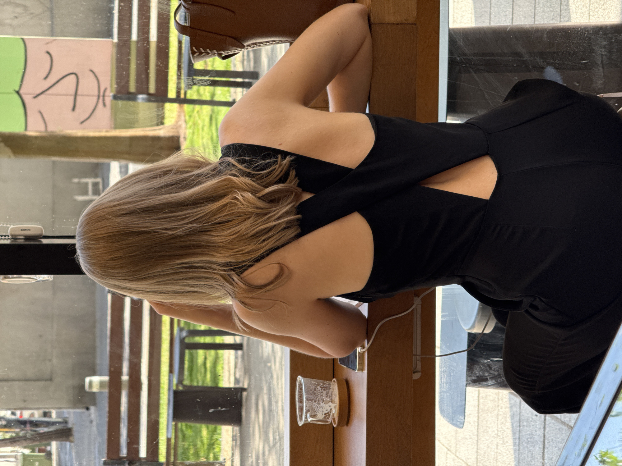 The image, oriented in portrait, captures a person from behind, seated at a light brown wooden table next to a large window. This setting suggests an indoor space, possibly a cafe or co-working area in Tel-Aviv, Israel.
The subject is a person with blonde or light brown hair, wearing a black top or dress with an open-back design featuring crisscrossing straps. Their arms are bent, resting on the table, and they appear to be looking forward, possibly out the window or at a device on the table. The skin on their back and arms is visible.
On the table, near the person, there's a clear glass with a wooden base, holding some residual liquid or foam. A smartphone, identified by its light-colored case and charging port, lies on the table connected to a white charging cable. Another small white rectangular device, possibly a power bank, is positioned near the window; the text "BANOG" is partially visible on it. A corner of a reflective screen, likely a laptop or tablet, is also visible on the lower right of the table.
Beyond the window, an outdoor scene unfolds under bright natural light, indicating it is daytime. Greenery, resembling grass and trees, can be seen, along with a wooden slatted fence or structure. In the background, there's a building with a light-colored wall featuring abstract black graffiti or art. The reflections on the glass suggest clear, possibly sunny weather.