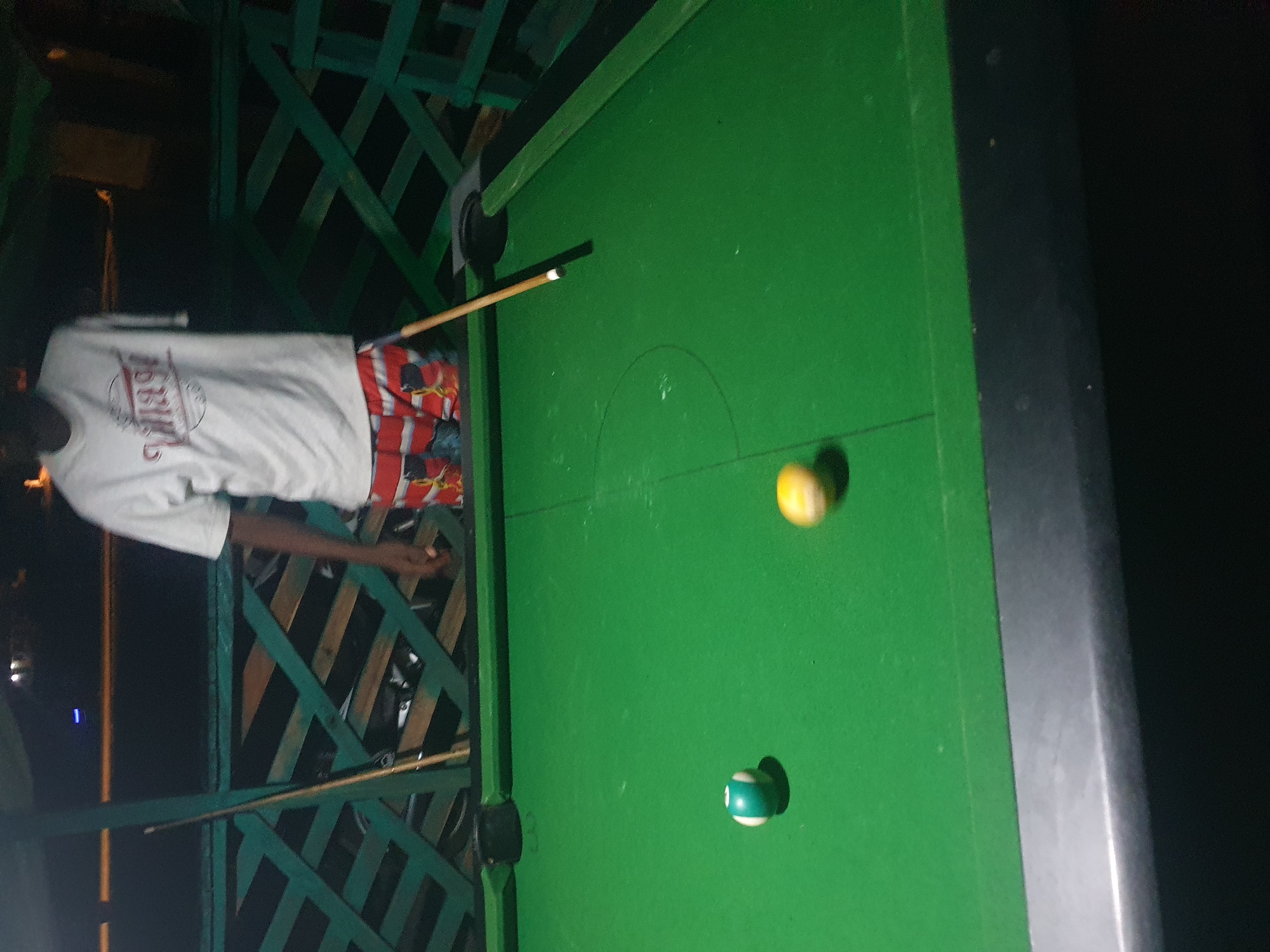A full-shot photograph captures a person playing pool at night, potentially in Lokoja, Nigeria, given the implied context. The scene is dimly lit, with the green felt of the pool table dominating the foreground. Two billiard balls are visible: one striped green and white, and another, a cue ball, appearing blurred as if in motion, possibly yellow or white. A cue stick rests on the table, angled across the surface, with its tip near a black pocket. Behind the pool table, a person stands wearing a white t-shirt with a circular graphic and red, white, and blue patterned shorts. Their right arm is not visible, suggesting it is out of frame or they have an amputation. The background features a green wooden lattice structure. The overall impression is one of casual nighttime recreation.