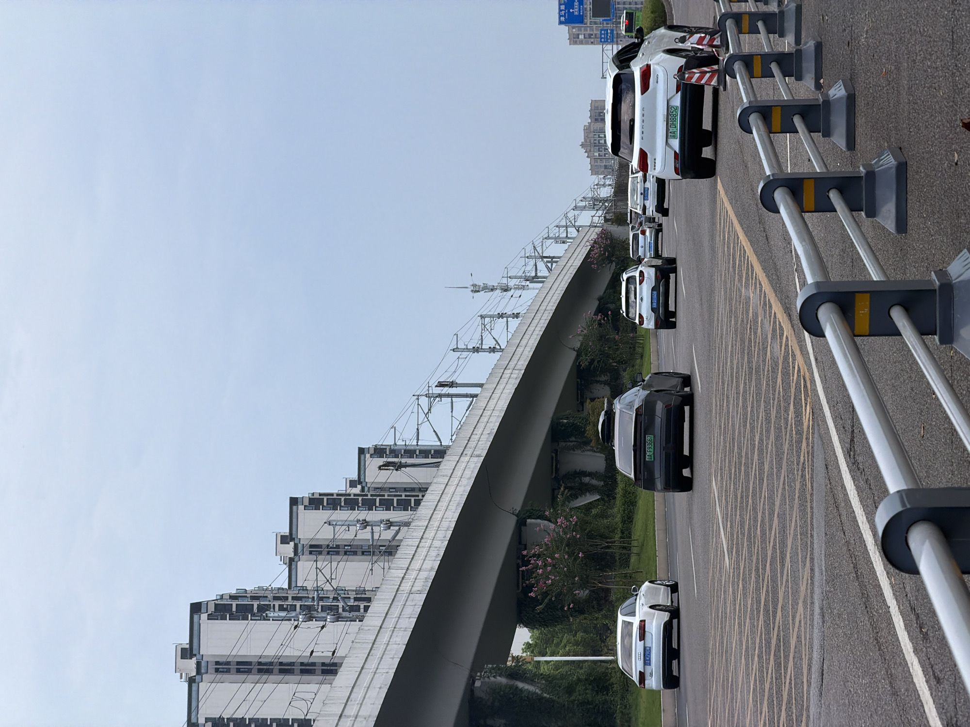 The image captures a street scene in Jiangjin, China, under an overcast sky. An elevated railway track structure dominates the upper portion of the frame, supported by concrete pillars. The track is lined with electrical infrastructure for trains. Beneath the elevated track, a road is visible with several vehicles in motion.
In the foreground, a dark gray car with a rooftop cargo box is driving away from the viewer. To its left, a white Audi sedan is also moving in the same direction. Further ahead, a white SUV is visible in the right lane, with a dark SUV and several other vehicles in the lanes to the left. The road is marked with yellow grid lines and white lane dividers.
On the left side of the road, greenery and some flowering trees are present. In the background, several tall, modern residential buildings are visible, aligned in a row. A blue traffic sign with Chinese characters is also visible on the right side of the road. The overall impression is of a busy urban transportation artery.